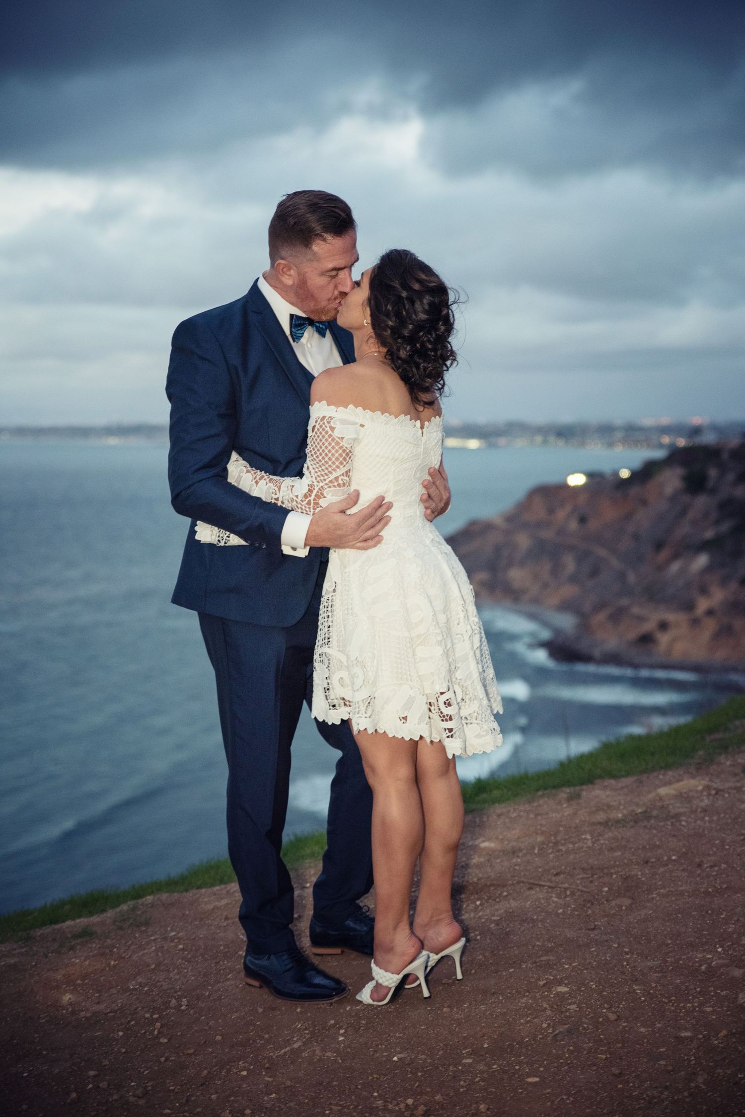A couple shares a romantic kiss on a coastal cliff overlooking stormy waters, with the bride in a short lace dress.