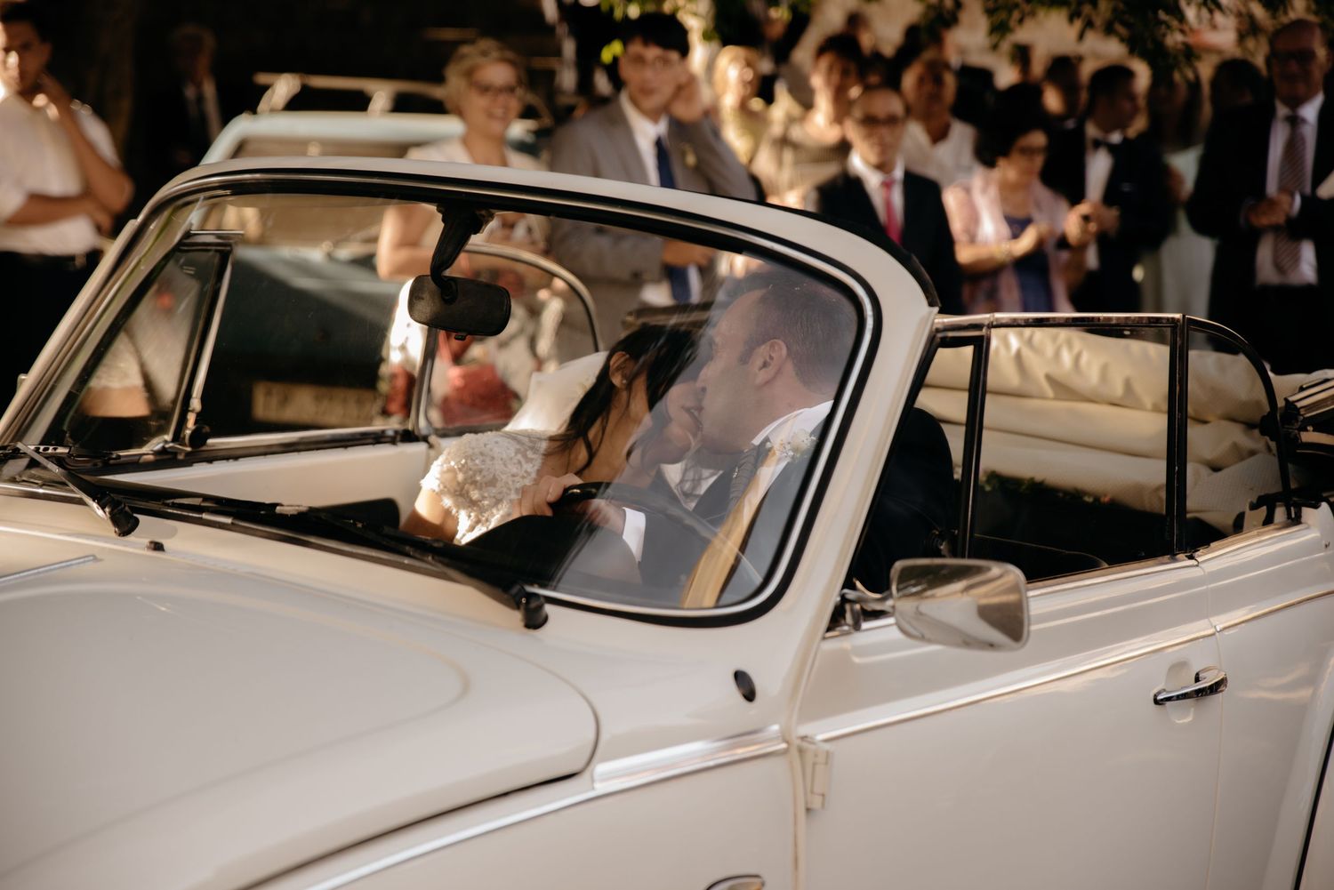 A classic white vintage car drives through a crowd during what appears to be a wedding celebration or special event.