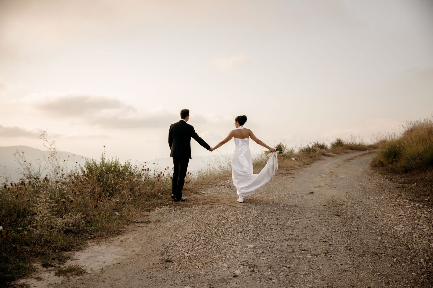 A couple in wedding attire walks along a dirt path at sunset holding hands while the bride's dress flows in the breeze.