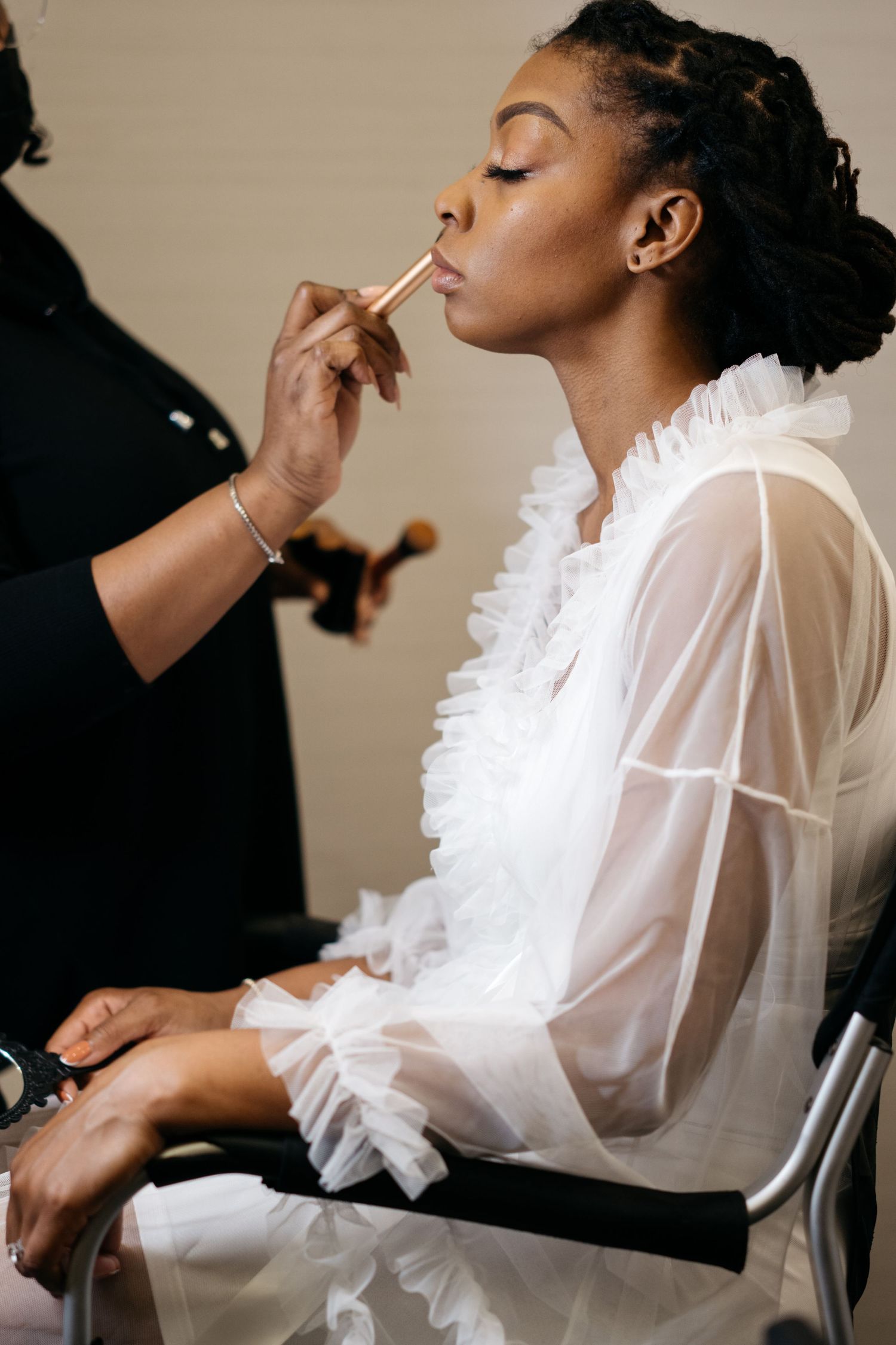 A makeup artist applies cosmetics to a model wearing an elegant white ruffled blouse during a professional beauty session.