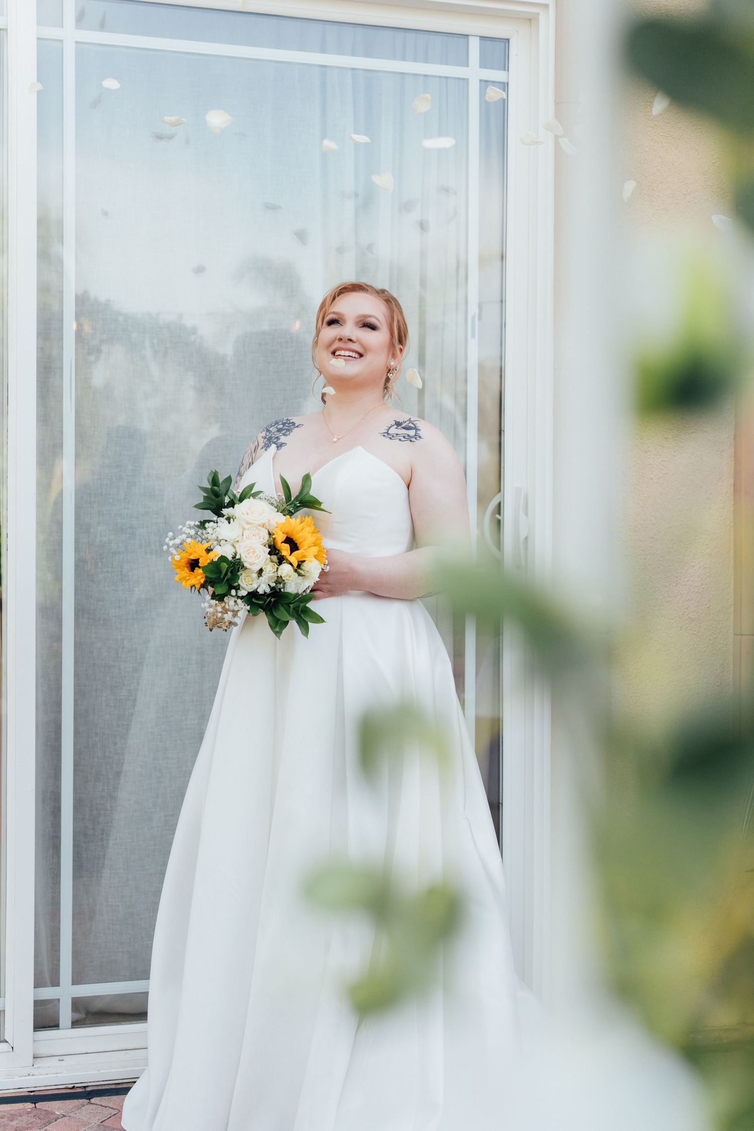 Bride in lace-sleeved gown holding yellow and white bouquet against window backdrop.