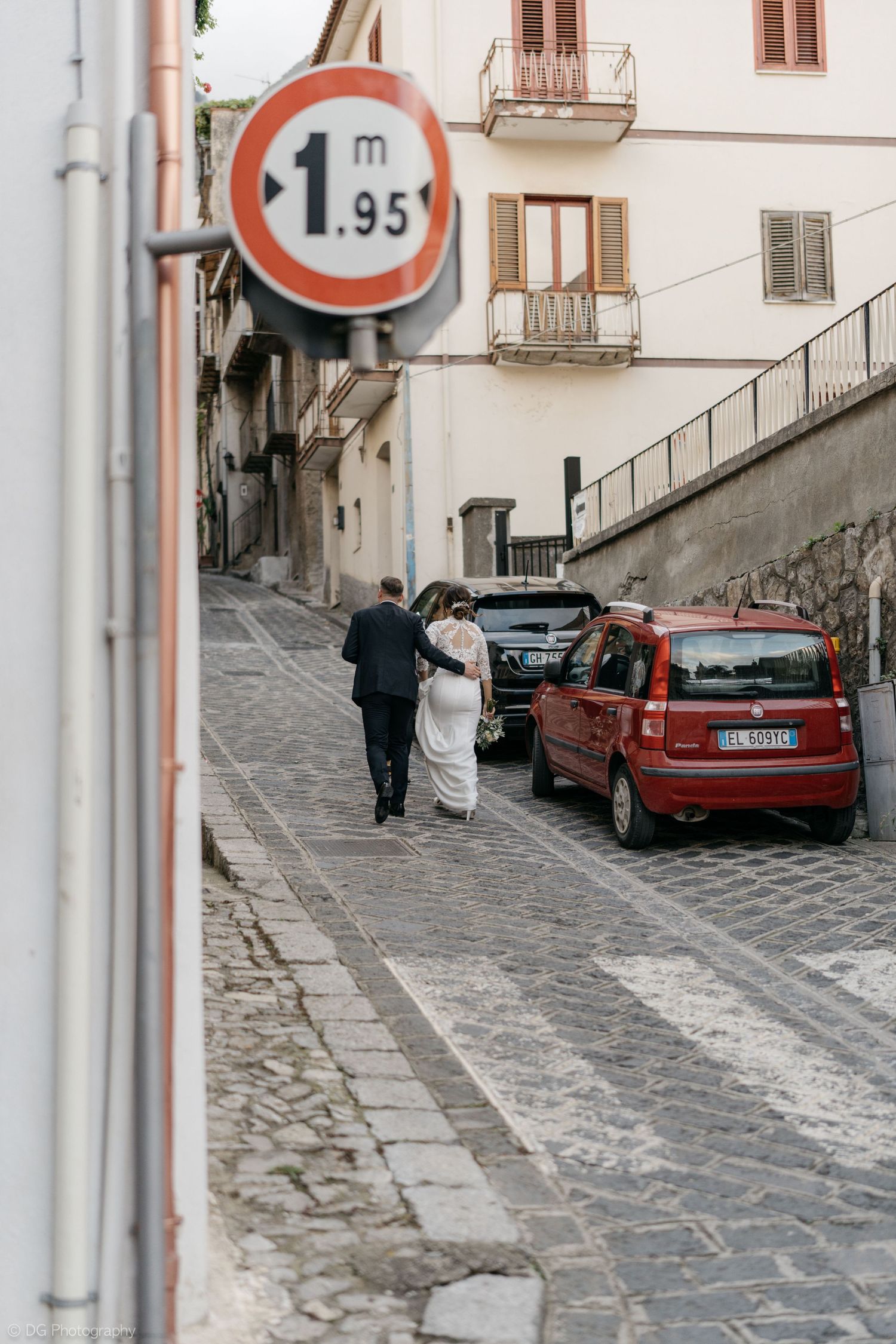 A narrow cobblestone street in Italy with a height restriction sign, parked cars, and two people walking uphill.