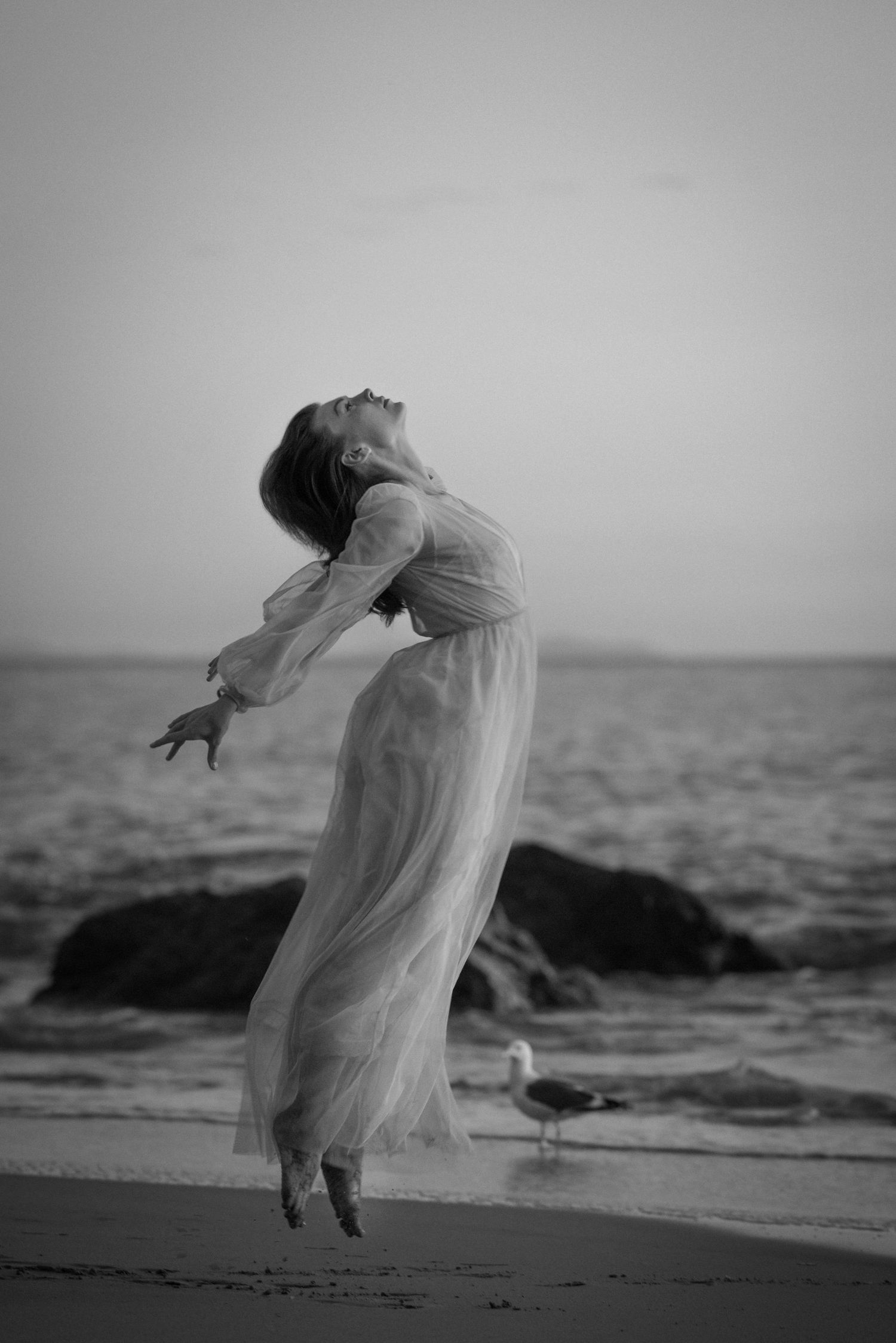 A graceful figure in a flowing white dress leaps ethereally on a beach at dusk with a seagull in the background.