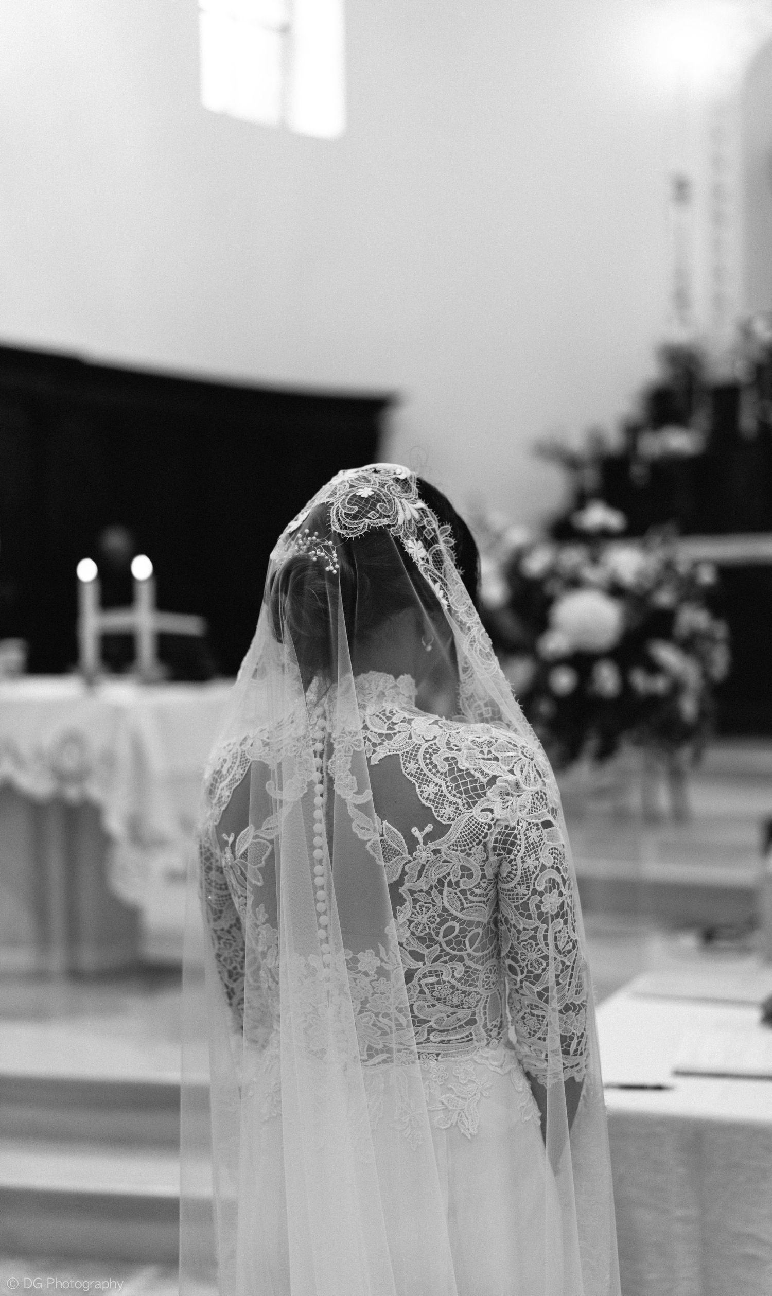 A bride wearing an elegant lace wedding dress and veil stands in a church during a ceremony, captured in black and white.