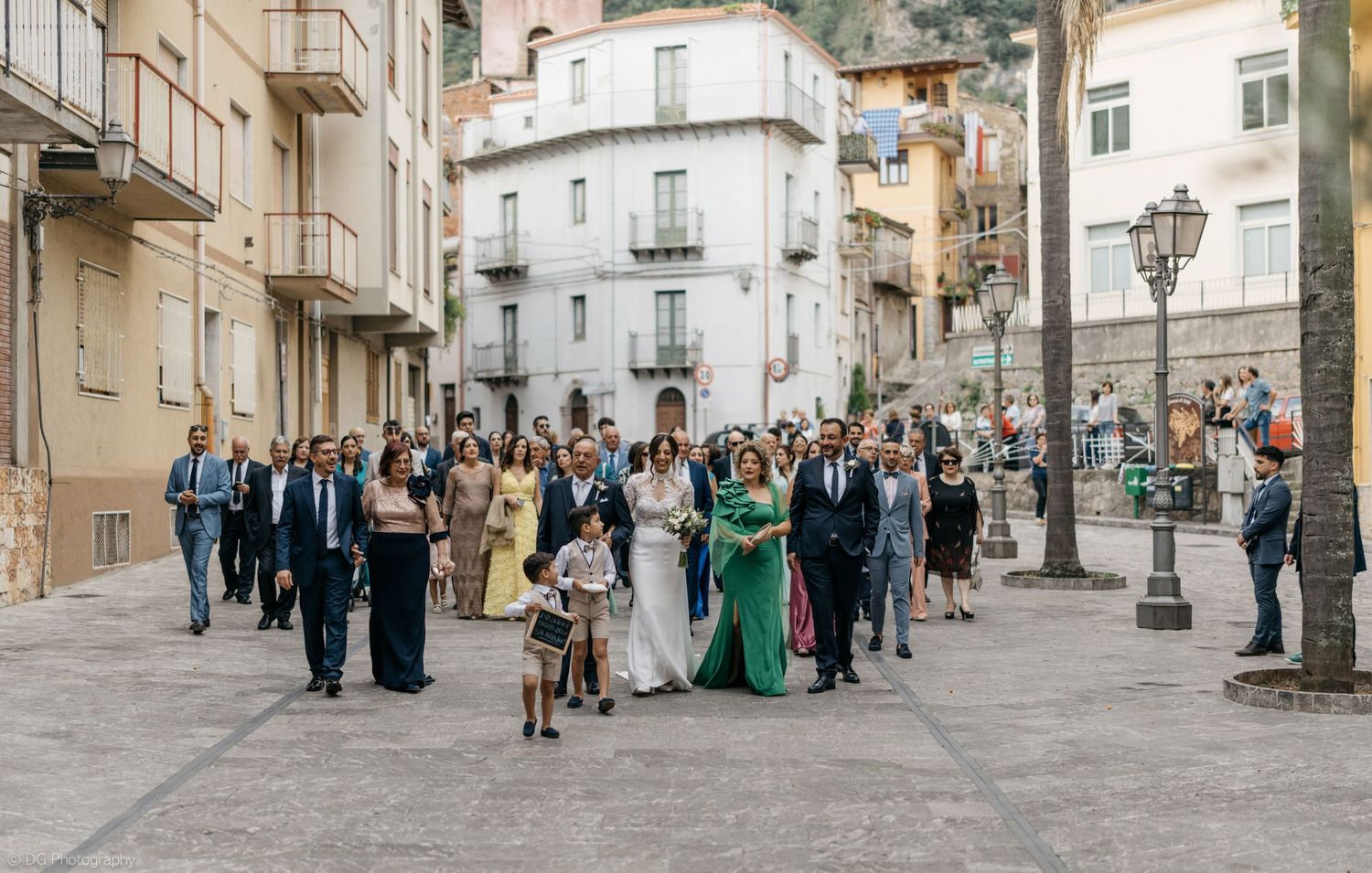 A panoramic view of a cobblestone street with people walking in formal attire outside historic buildings.