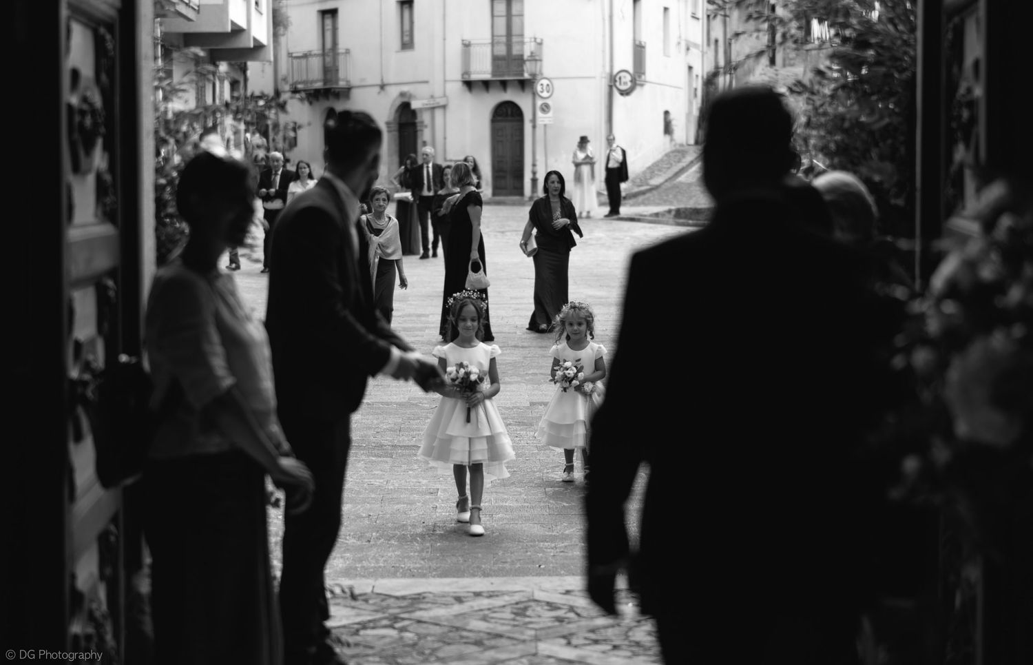 A black and white street scene in an old European town with figures walking along a cobblestone path.