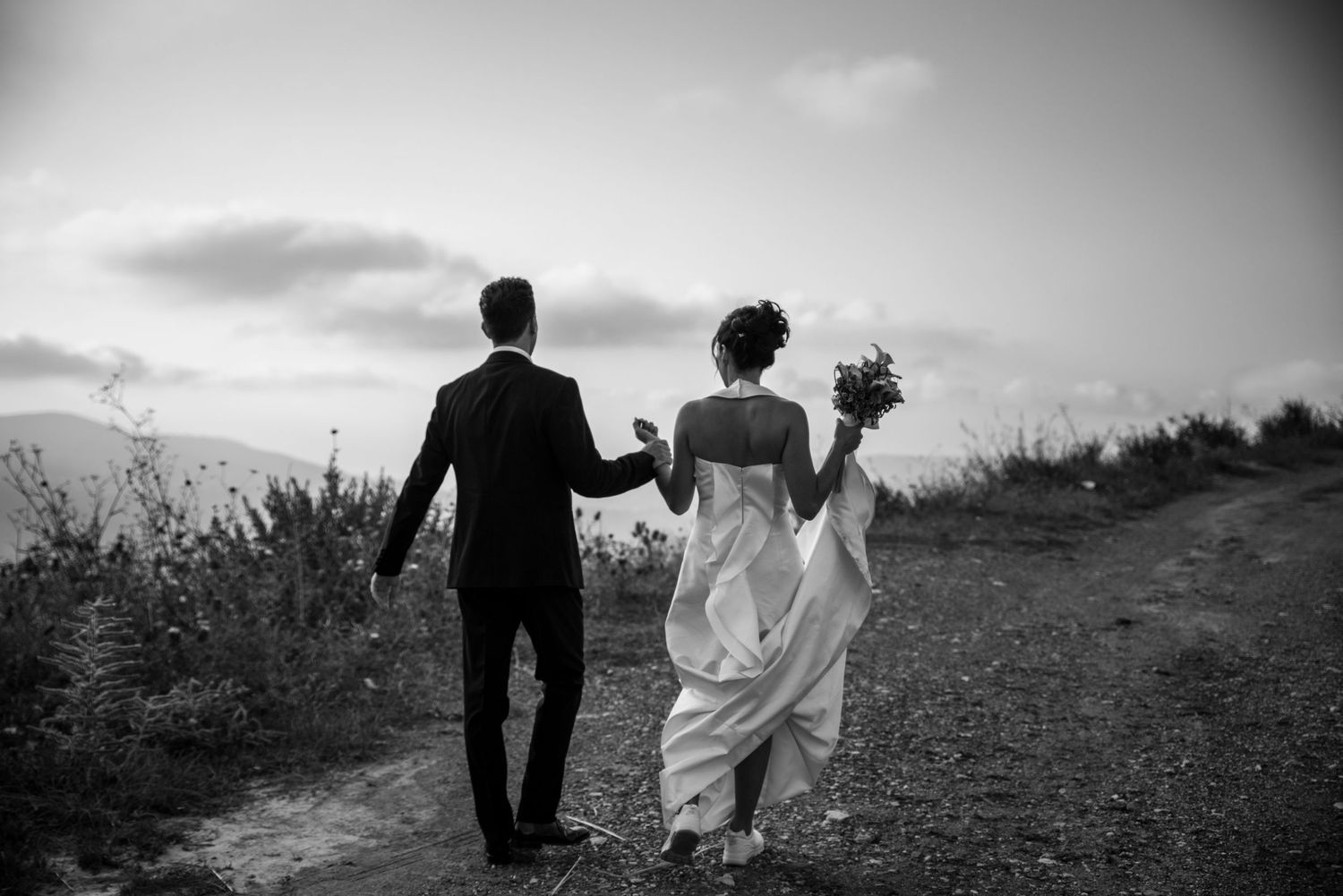 A couple walks hand in hand through a scenic field at sunset, their silhouettes creating a romantic black and white scene.