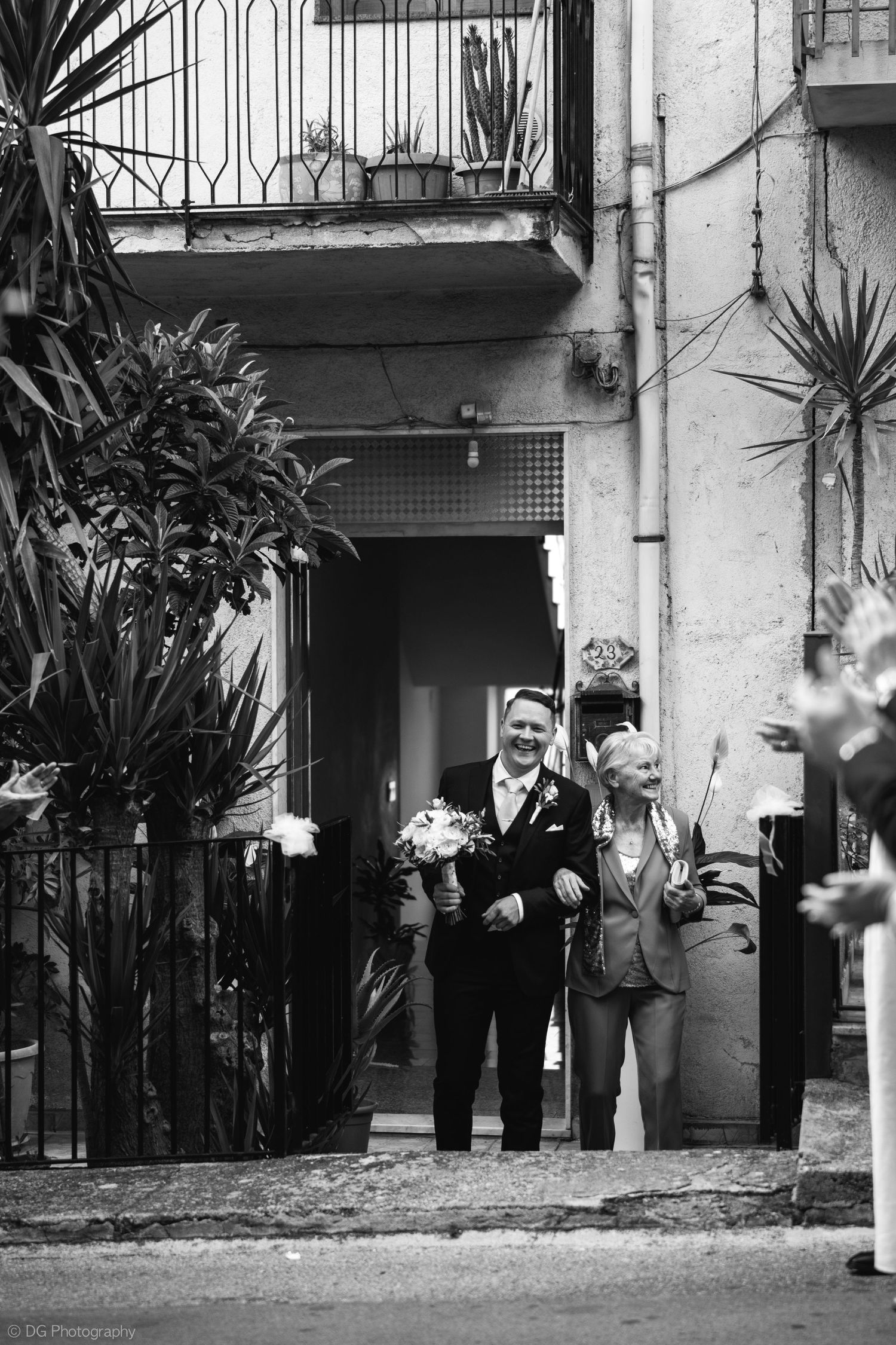 A black and white photo shows a group of people standing outside a building entrance with tropical plants.