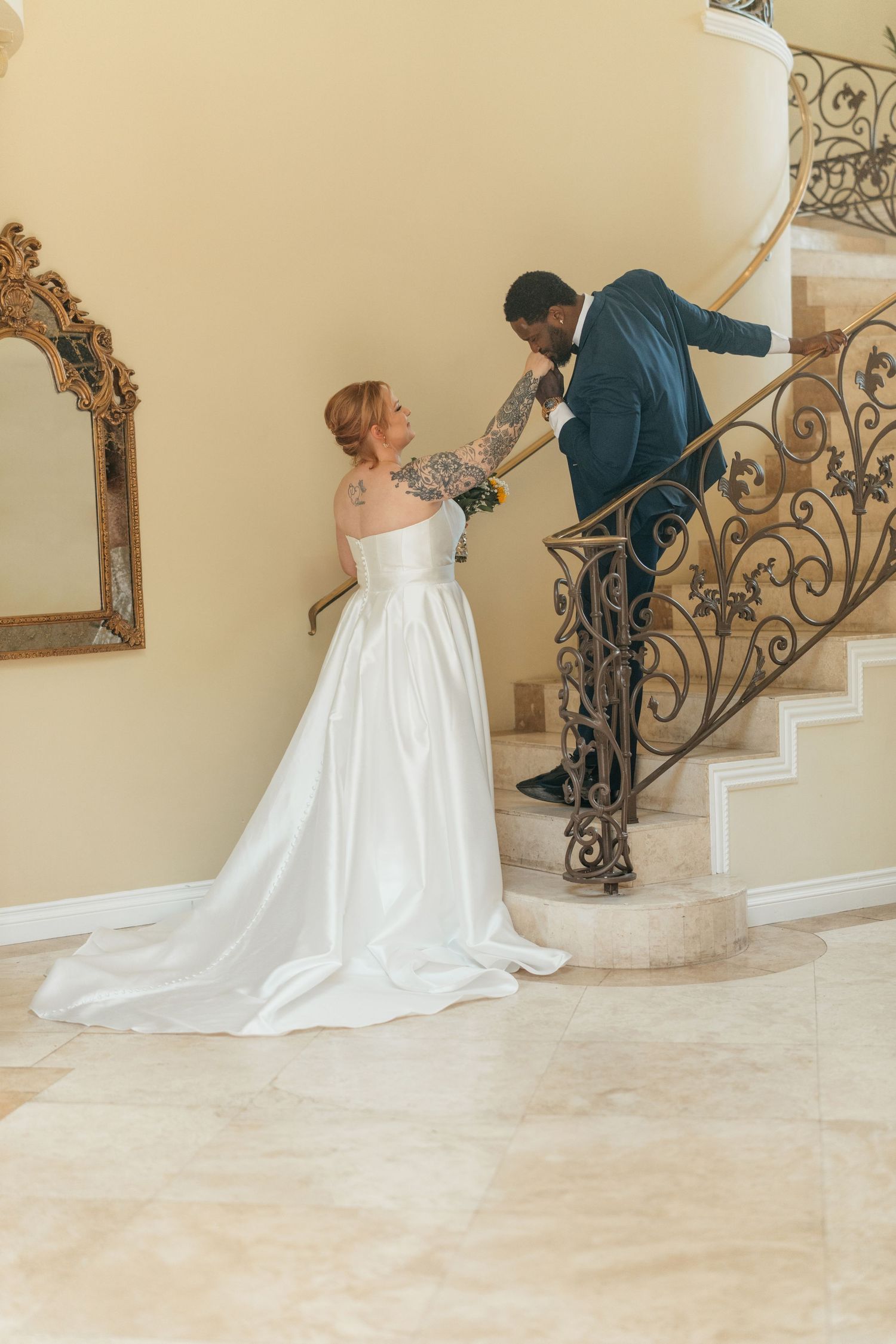 Bride in flowing white gown posing on elegant staircase with ornate iron railings.