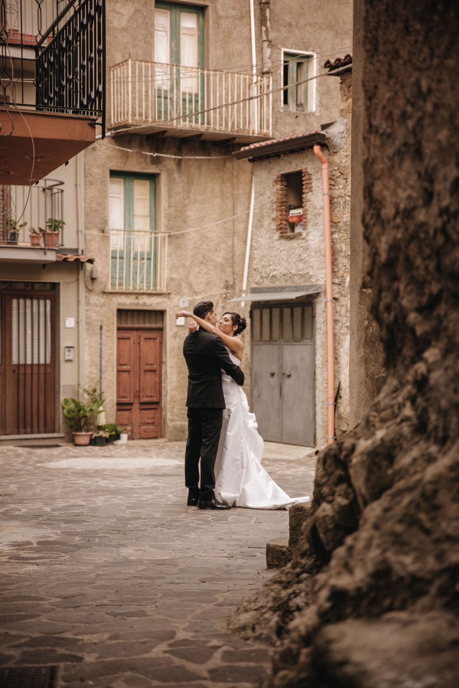 A romantic couple in wedding attire dances together on a cobblestone street in front of rustic European buildings.