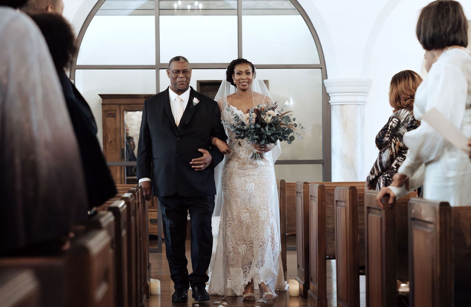 A couple walks down the aisle of a church with wooden pews and arched windows during their wedding ceremony.
