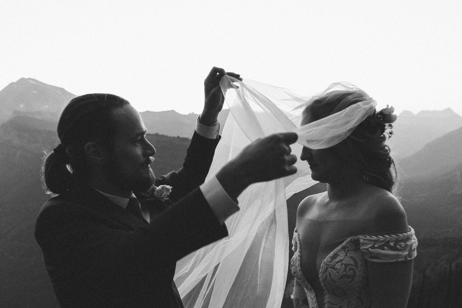 Black and white photo of a couple in wedding attire embracing with mountains in the background.