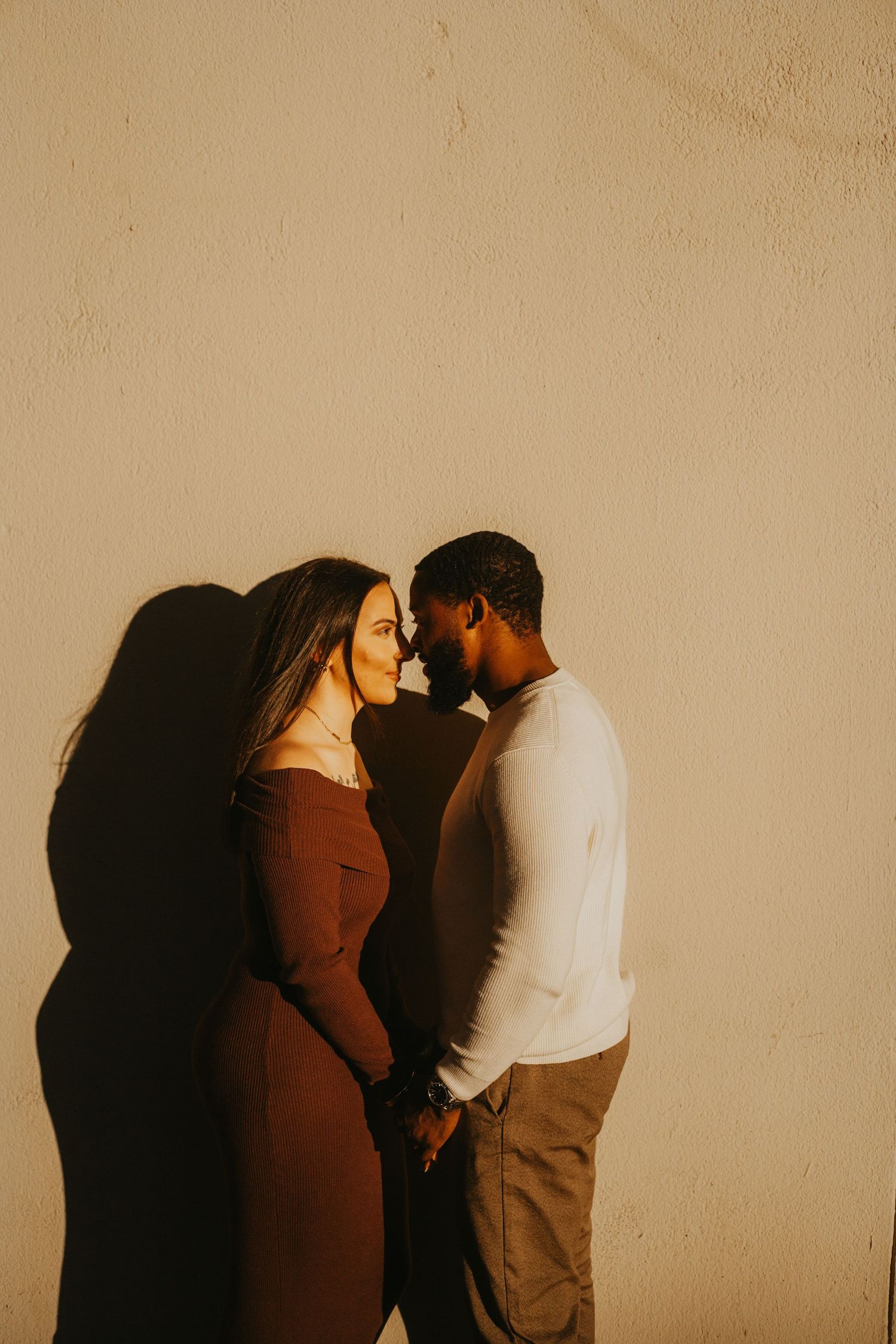 A couple in warm lighting poses against a beige wall for an intimate portrait.