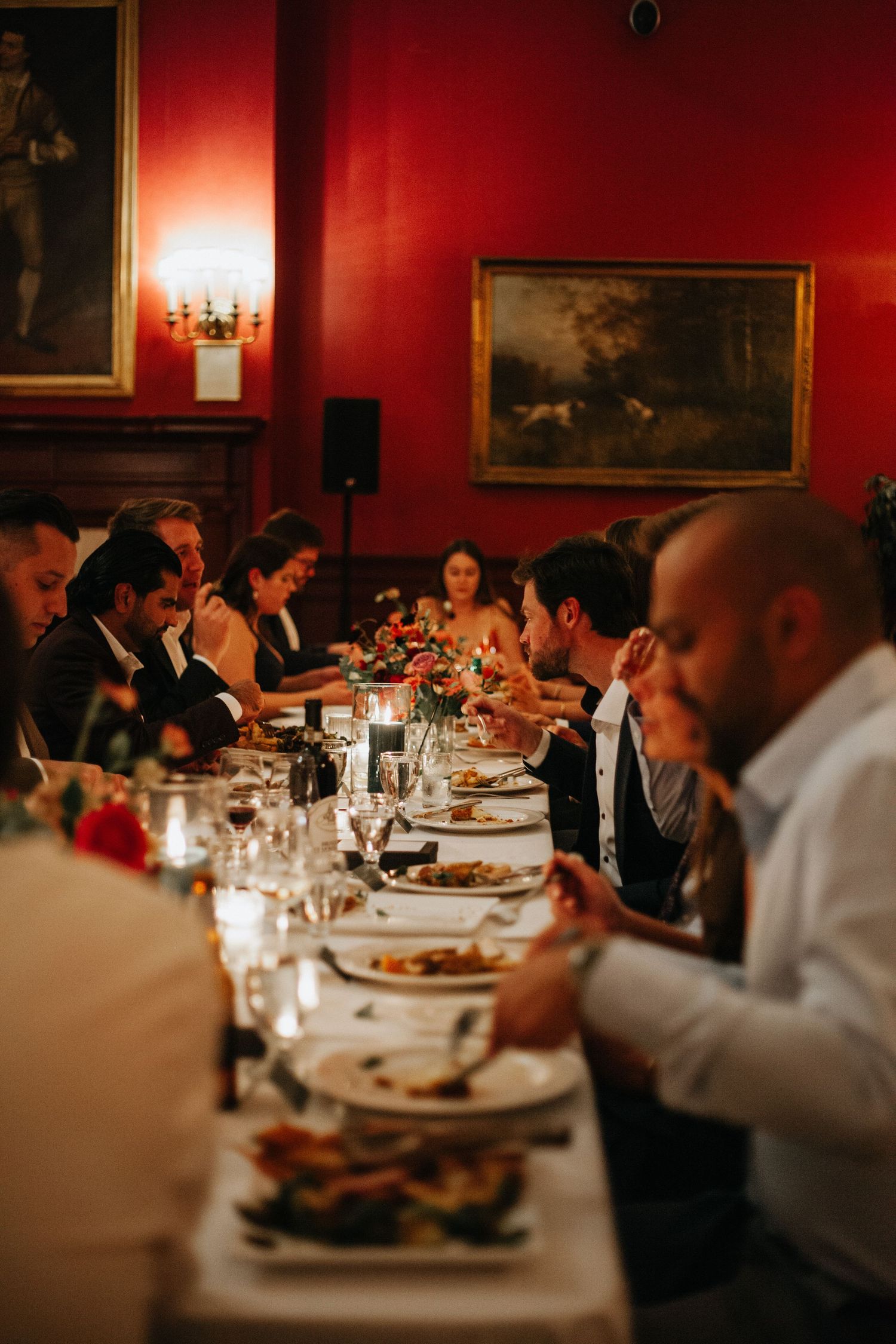 Wedding guests enjoying an elegant dinner at a long table in a formal dining room with red walls and vintage artwork.