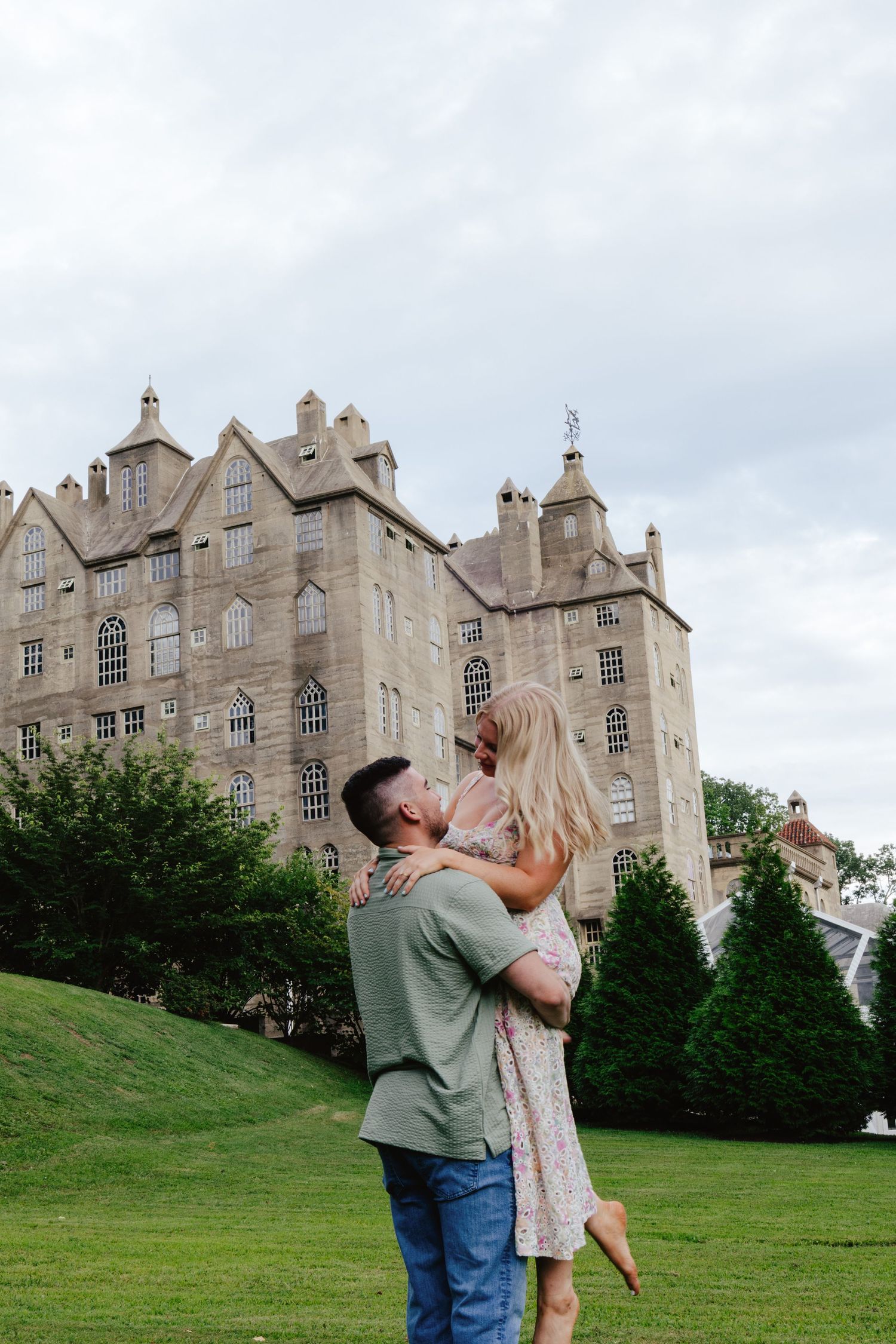 A couple embraces romantically in front of a grand stone castle-like building with Gothic architecture.