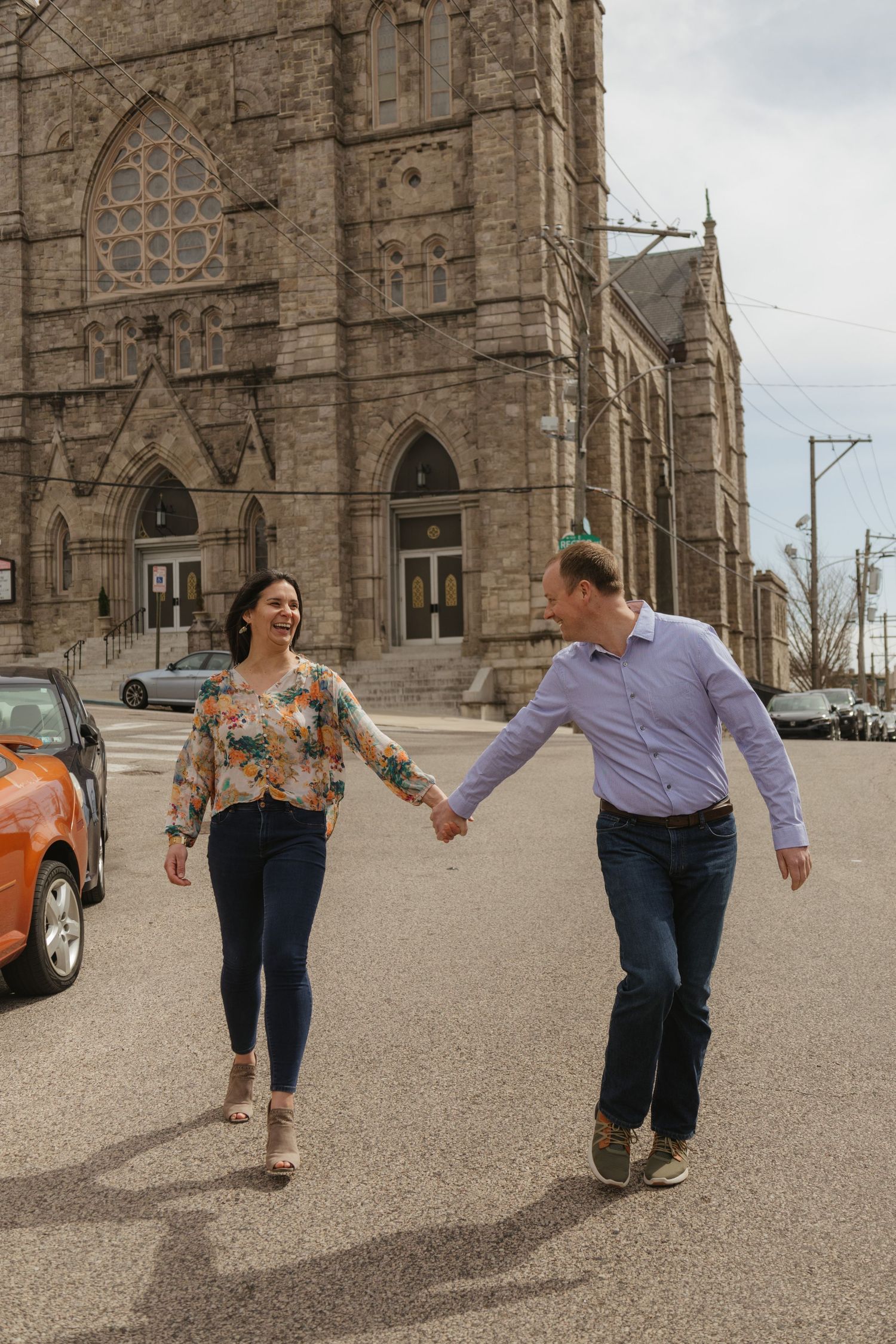 Couple holding hands while walking in front of a historic Gothic church building with orange car in background.