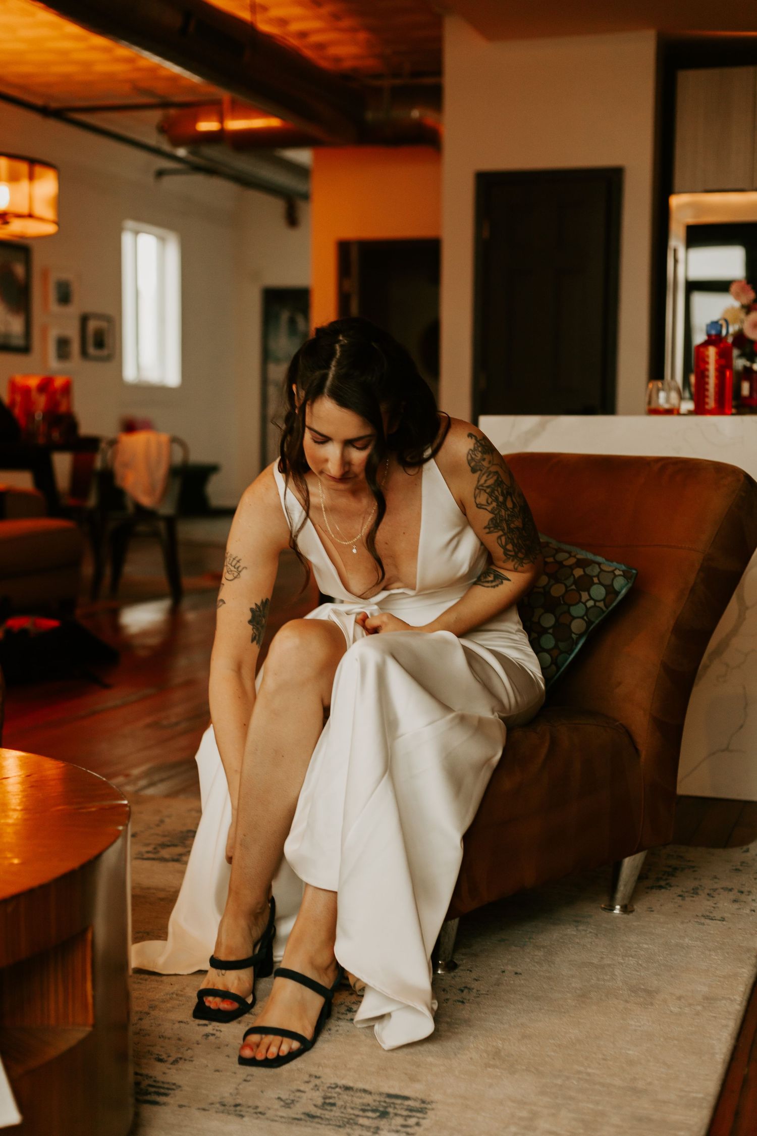 A bride in a white dress and tattoos adjusts her gown while sitting on a brown leather couch.