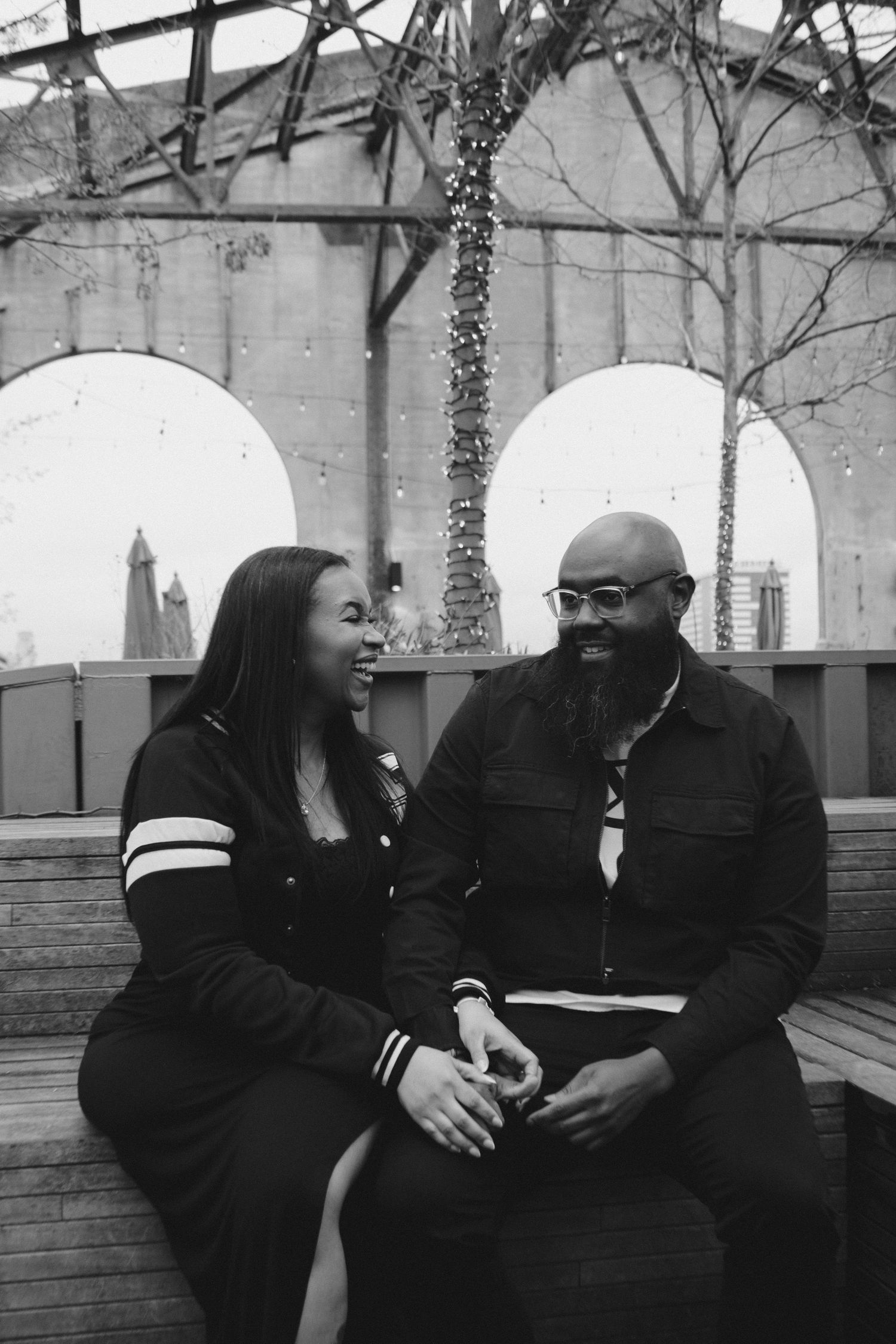 A couple shares a candid laughing moment on a rooftop deck with urban architecture in background.