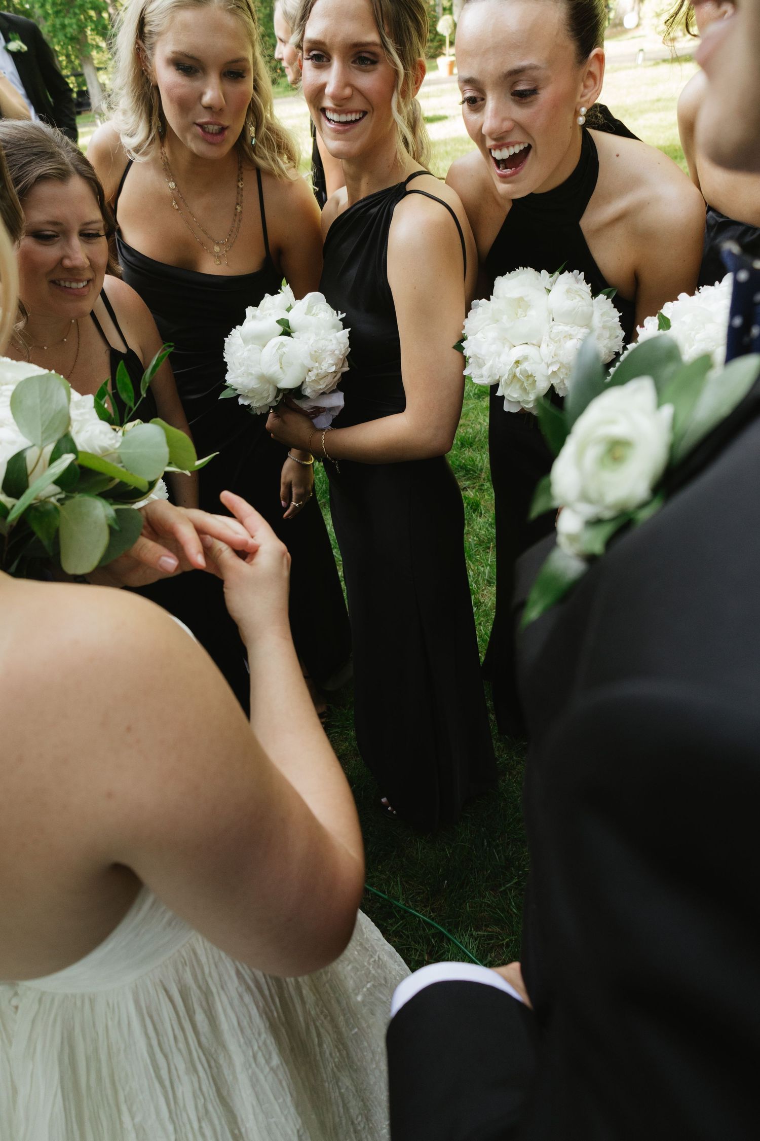A wedding party group in black dresses and white bouquets share a candid joyful moment outdoors.