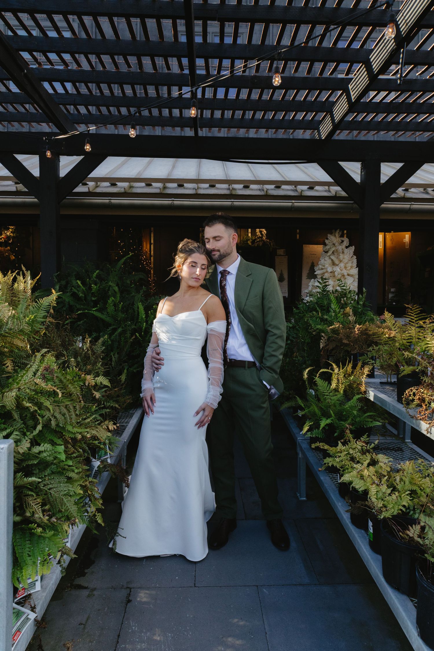 A couple in wedding attire poses together under a pergola with lush greenery in the background.