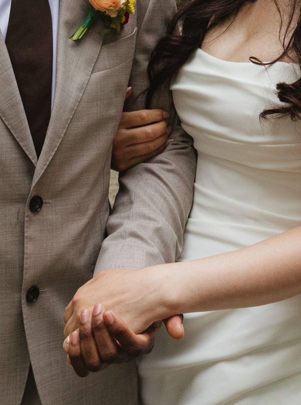 A couple holds hands during their wedding, showing details of a gray suit with orange boutonniere and white bridal gown.