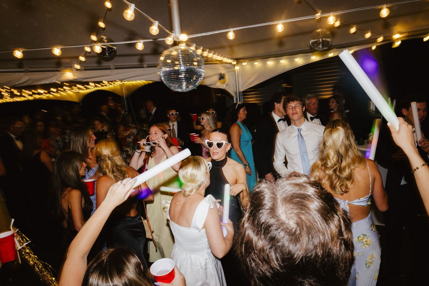 Wedding reception party with string lights and disco ball as guests dance with glow sticks.