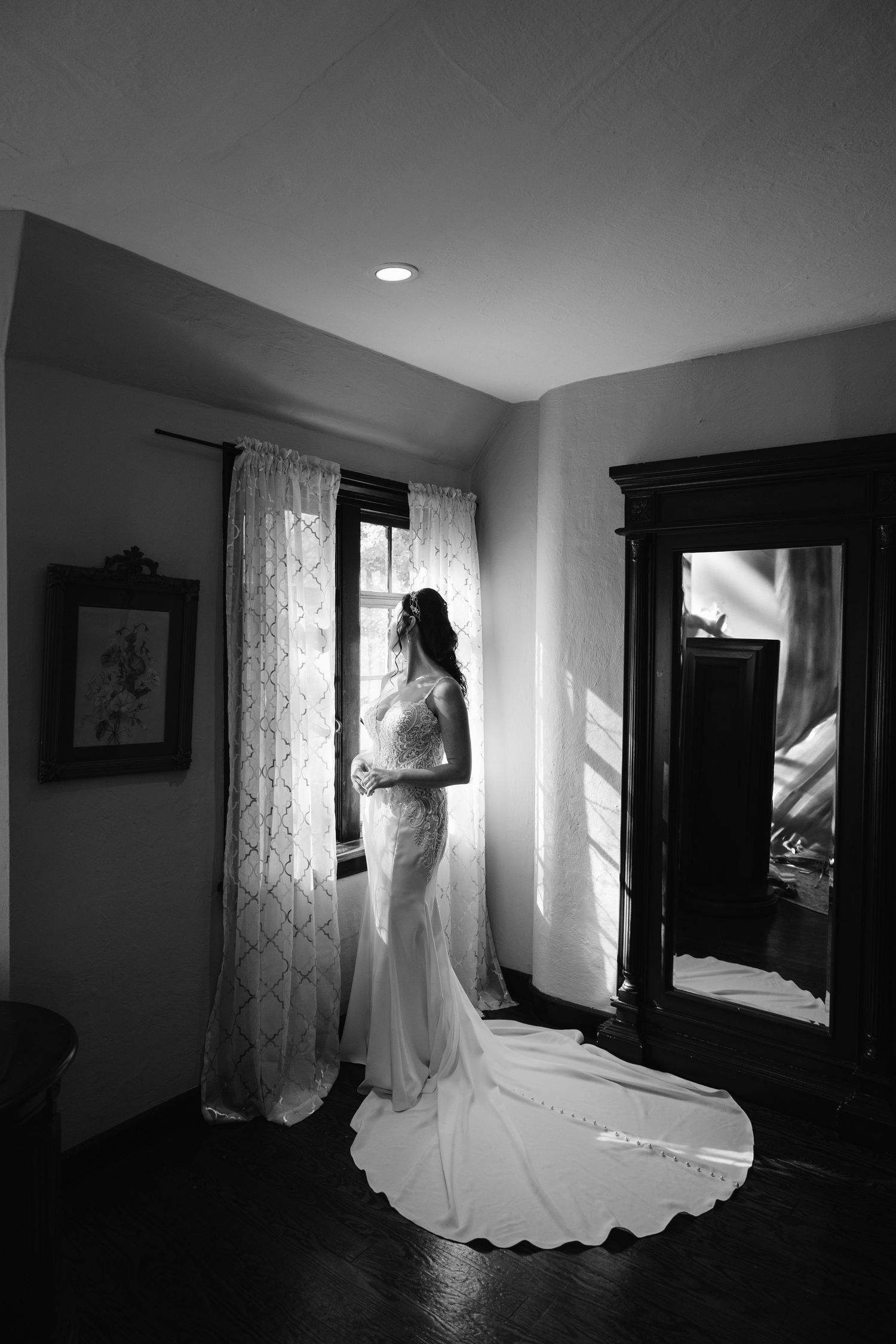 A dramatic black and white silhouette of a bride in her wedding gown standing by a window in a sunlit room.