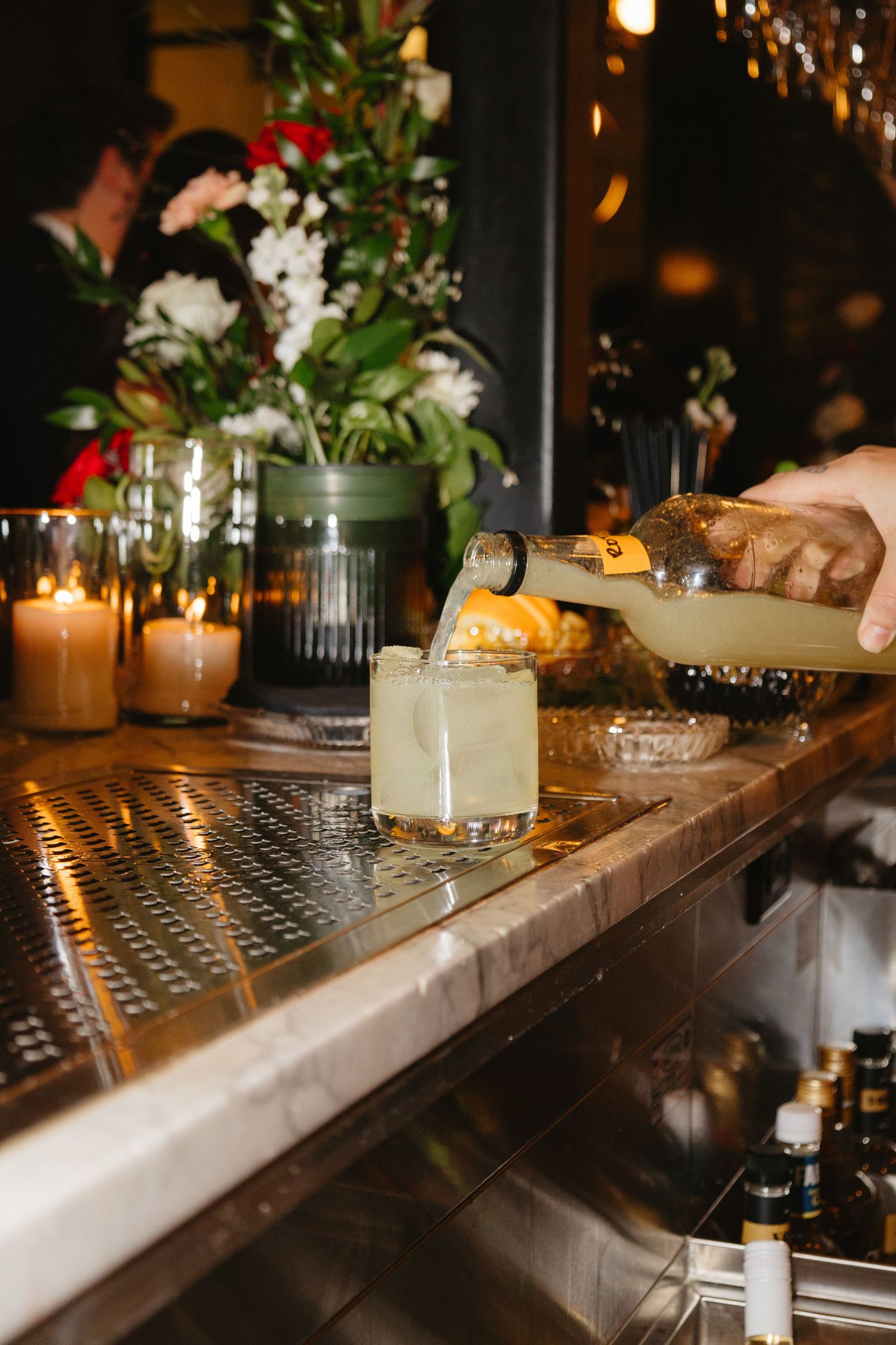 Bartender pouring crafted cocktail at elegant bar with floral arrangements and candles.