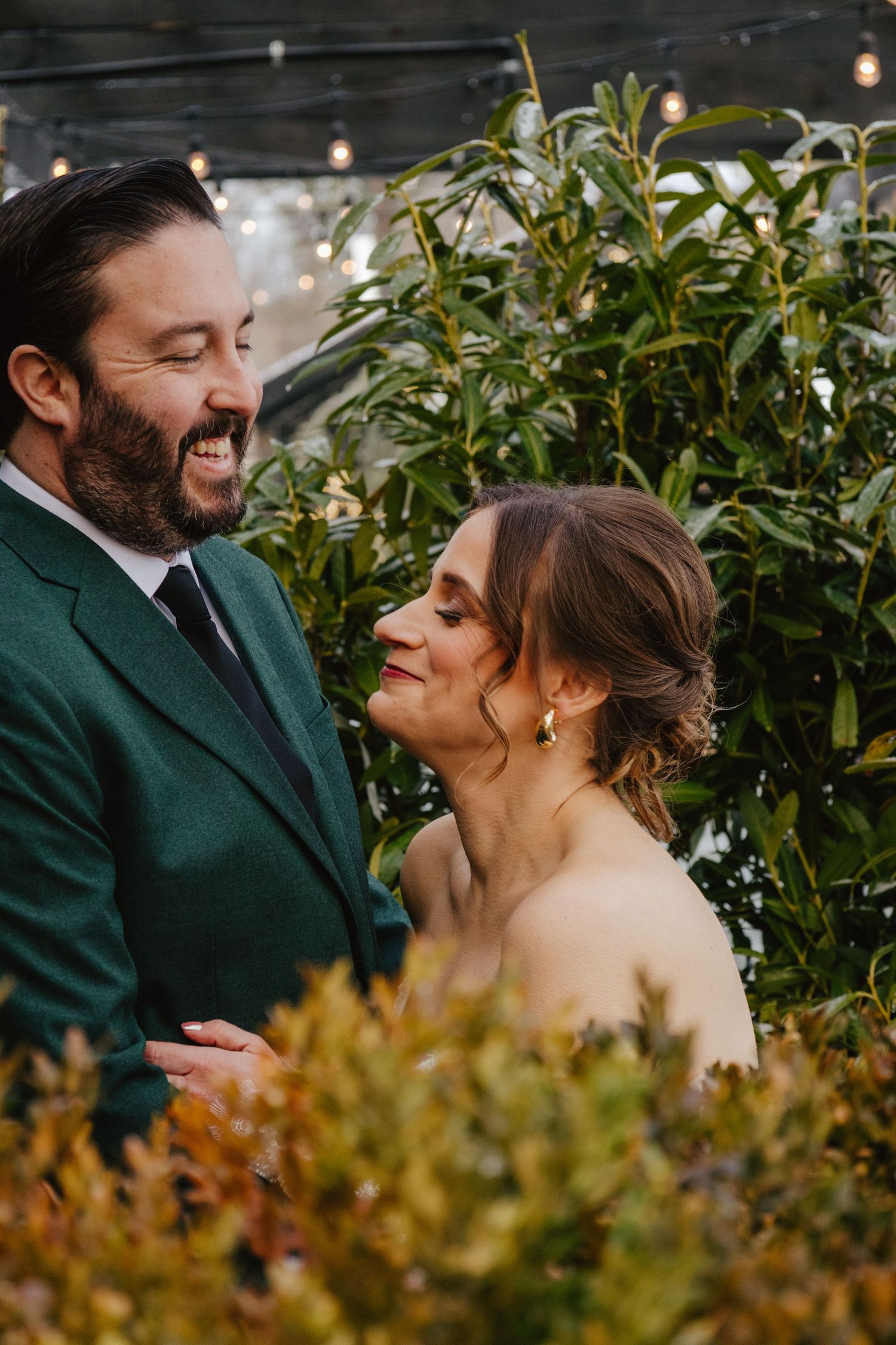 A couple shares a joyful moment surrounded by lush greenery at an outdoor wedding celebration.