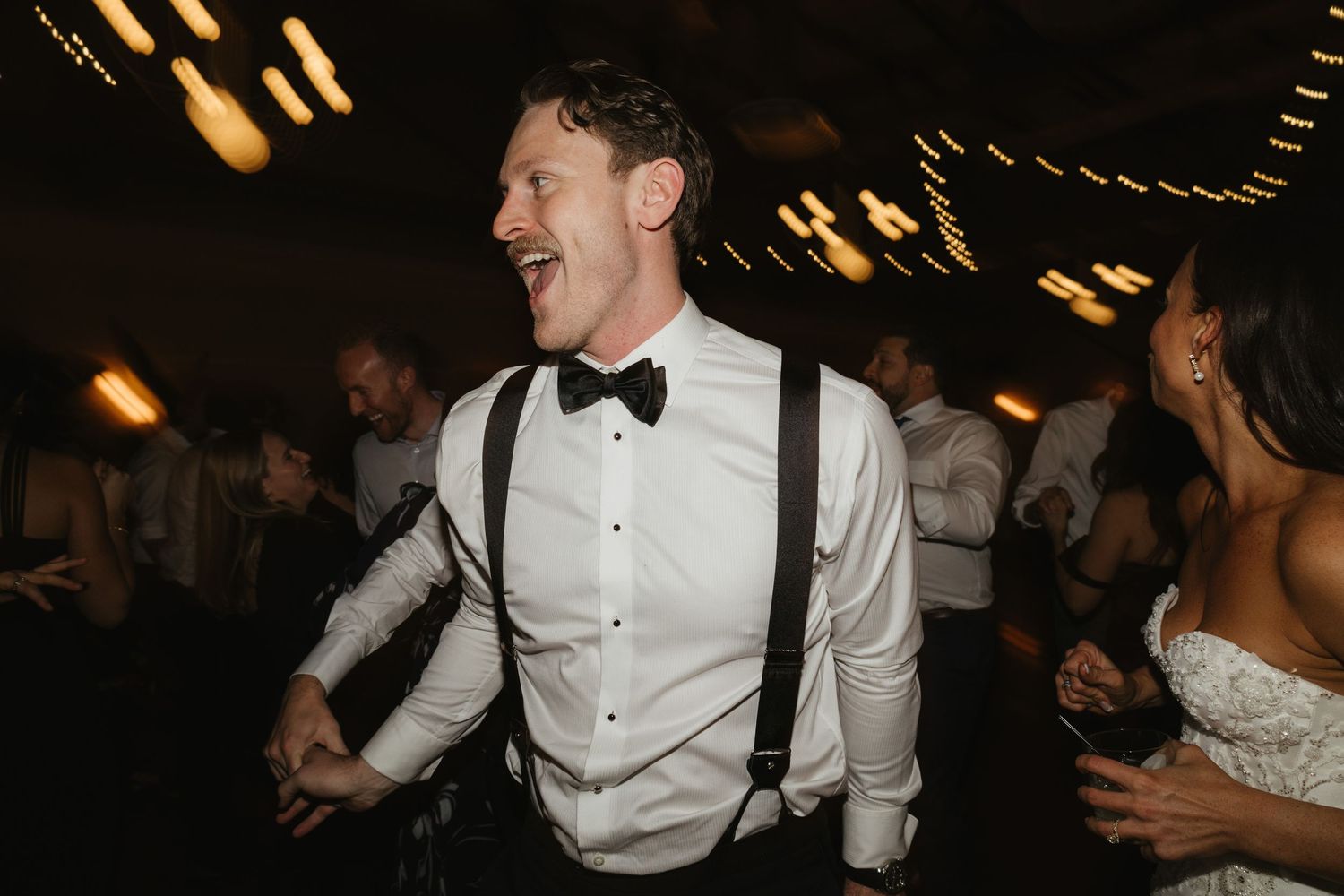 A person in suspenders and bow tie dances enthusiastically at a reception celebration.
