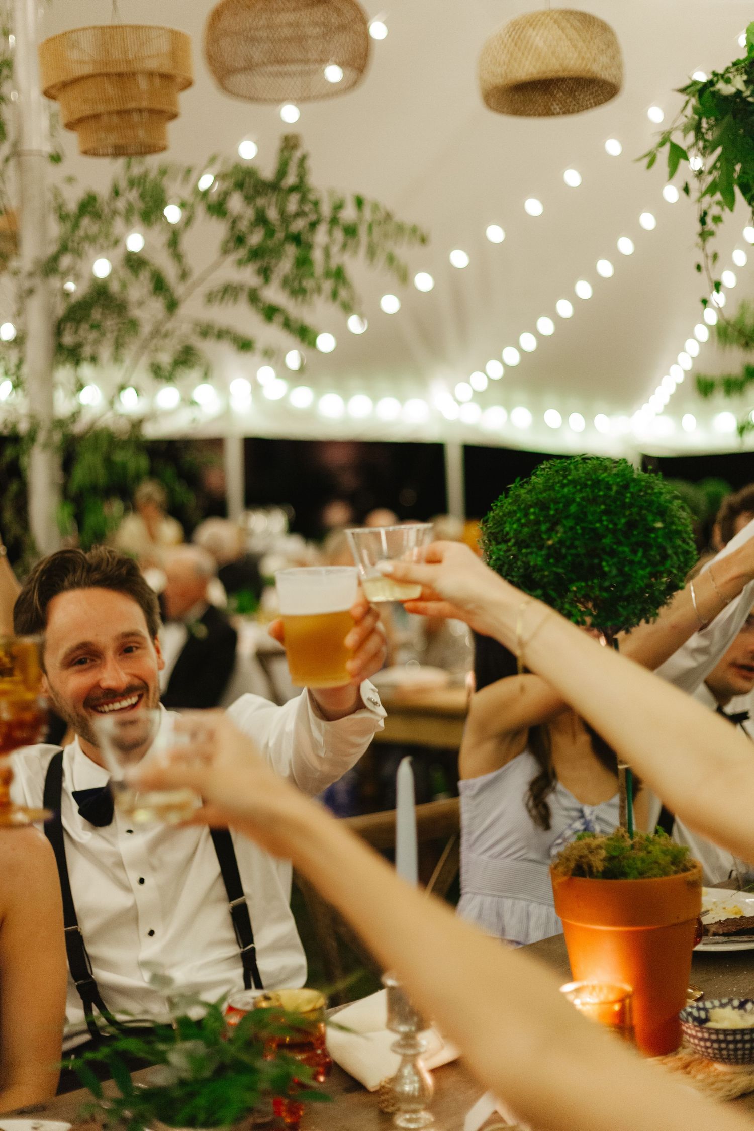 Friends raising glasses in a toast at an outdoor party under string lights with wicker lamps overhead.