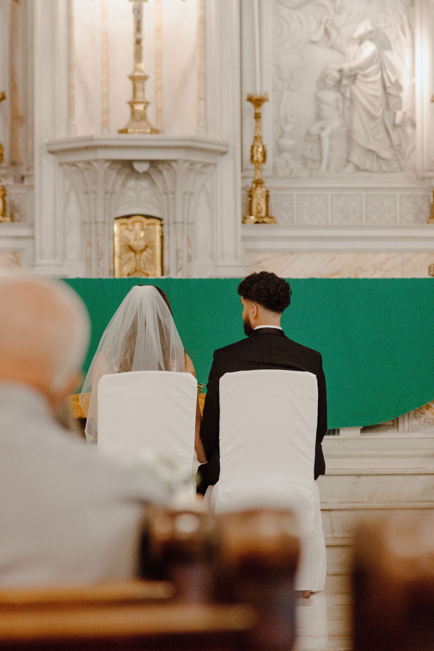 A couple seated at the altar during their wedding ceremony with ornate church decorations in the background.