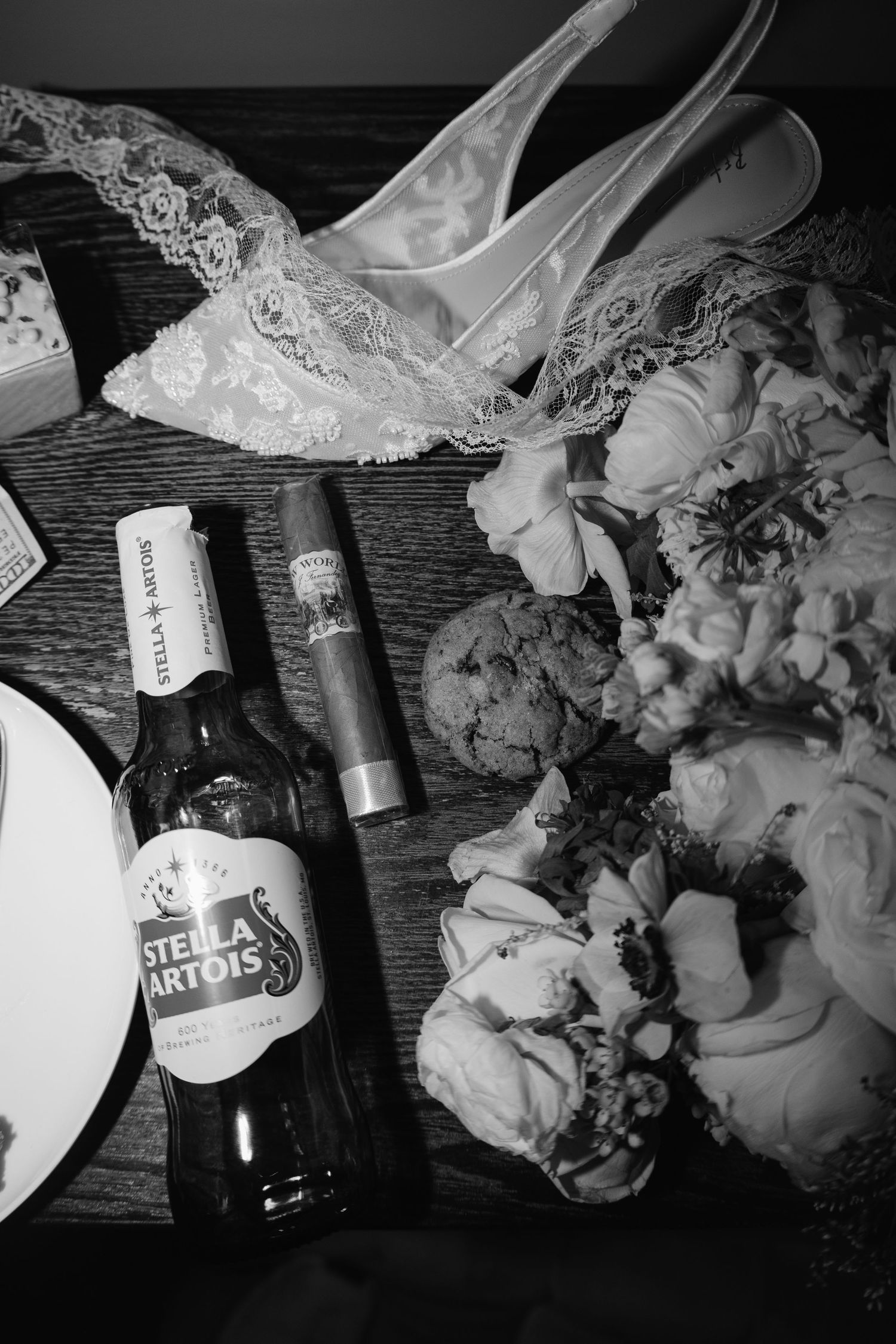 Wedding details including lace shoes, flowers and Stella Artois beer bottle on wooden surface.