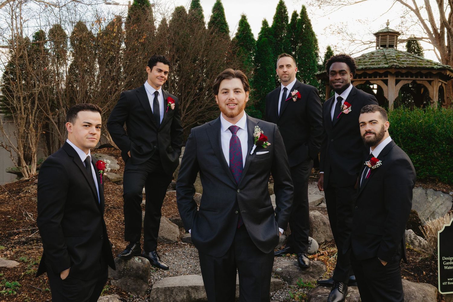 Wedding party in black suits and red ties pose together outdoors during autumn with trees in the background.
