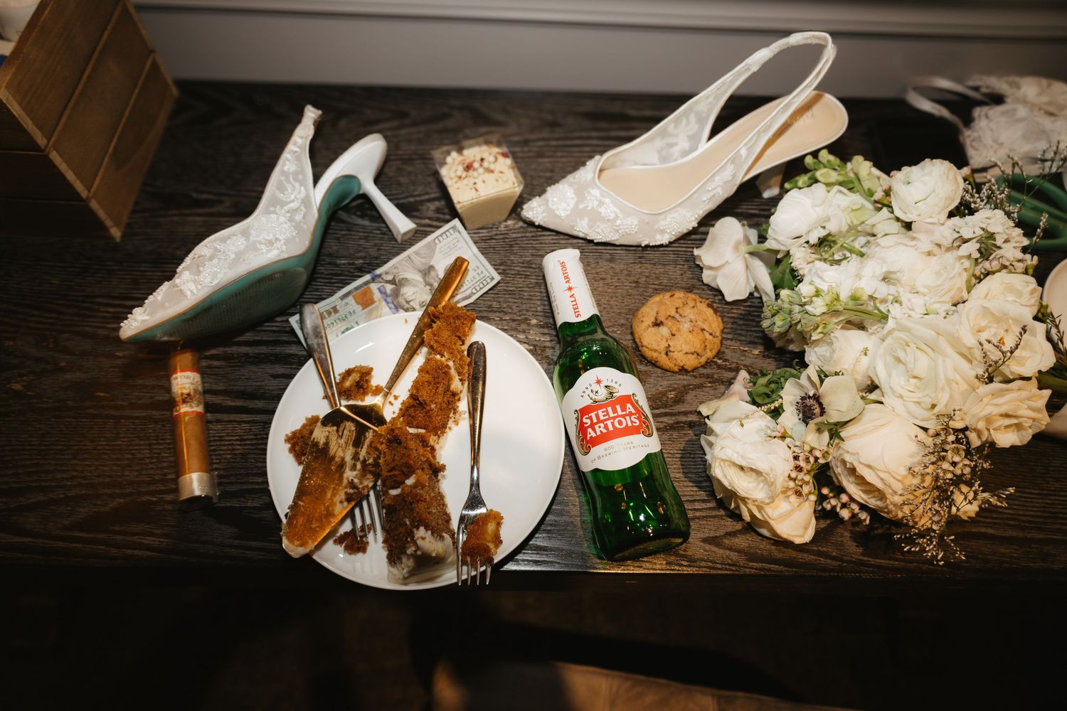 Wedding reception details featuring bridal shoes, flowers, and refreshments on dark wooden table.