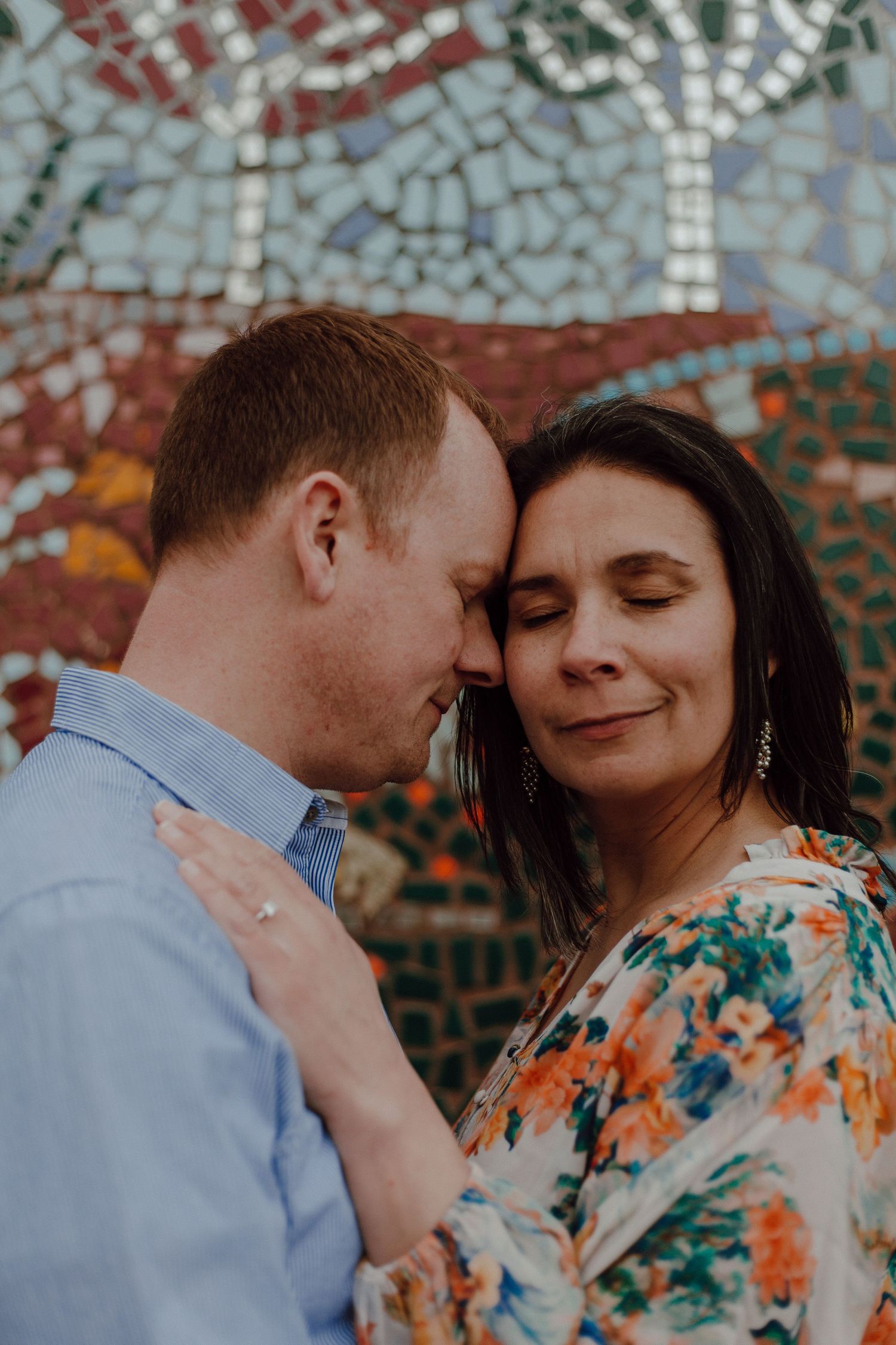 Romantic couple embracing in front of colorful mosaic wall artwork.