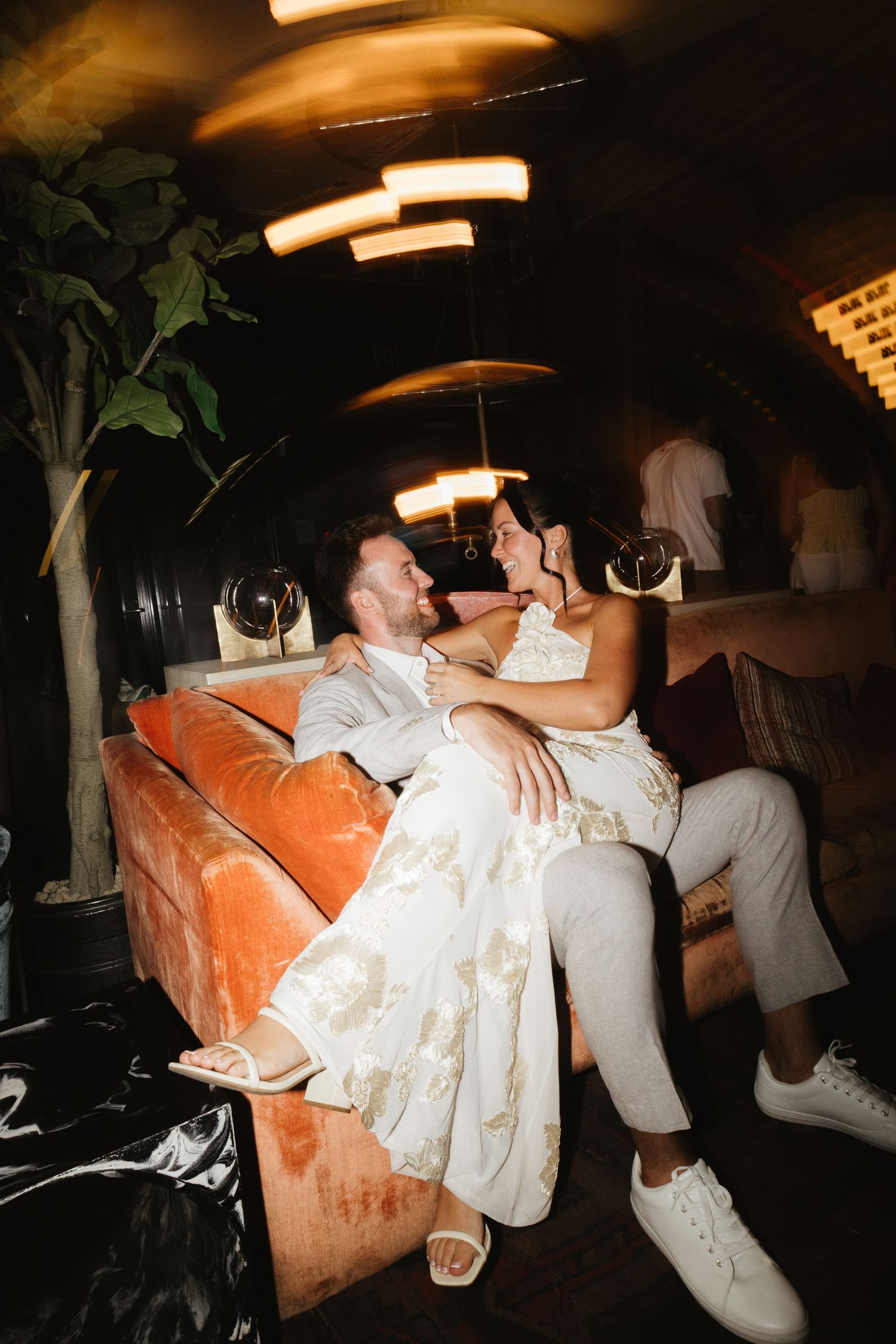 Two people in white outfits share a romantic moment on an orange couch in a dimly lit room.