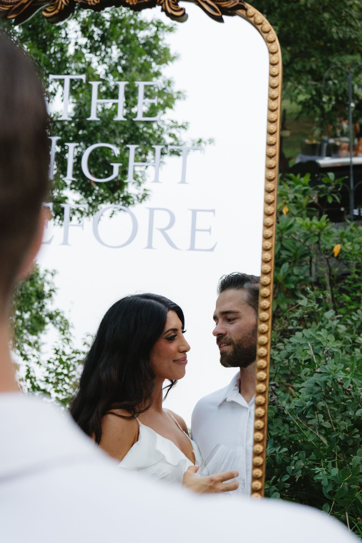 A couple poses lovingly together in an outdoor garden setting with an ornate gold mirror frame and greenery in background.
