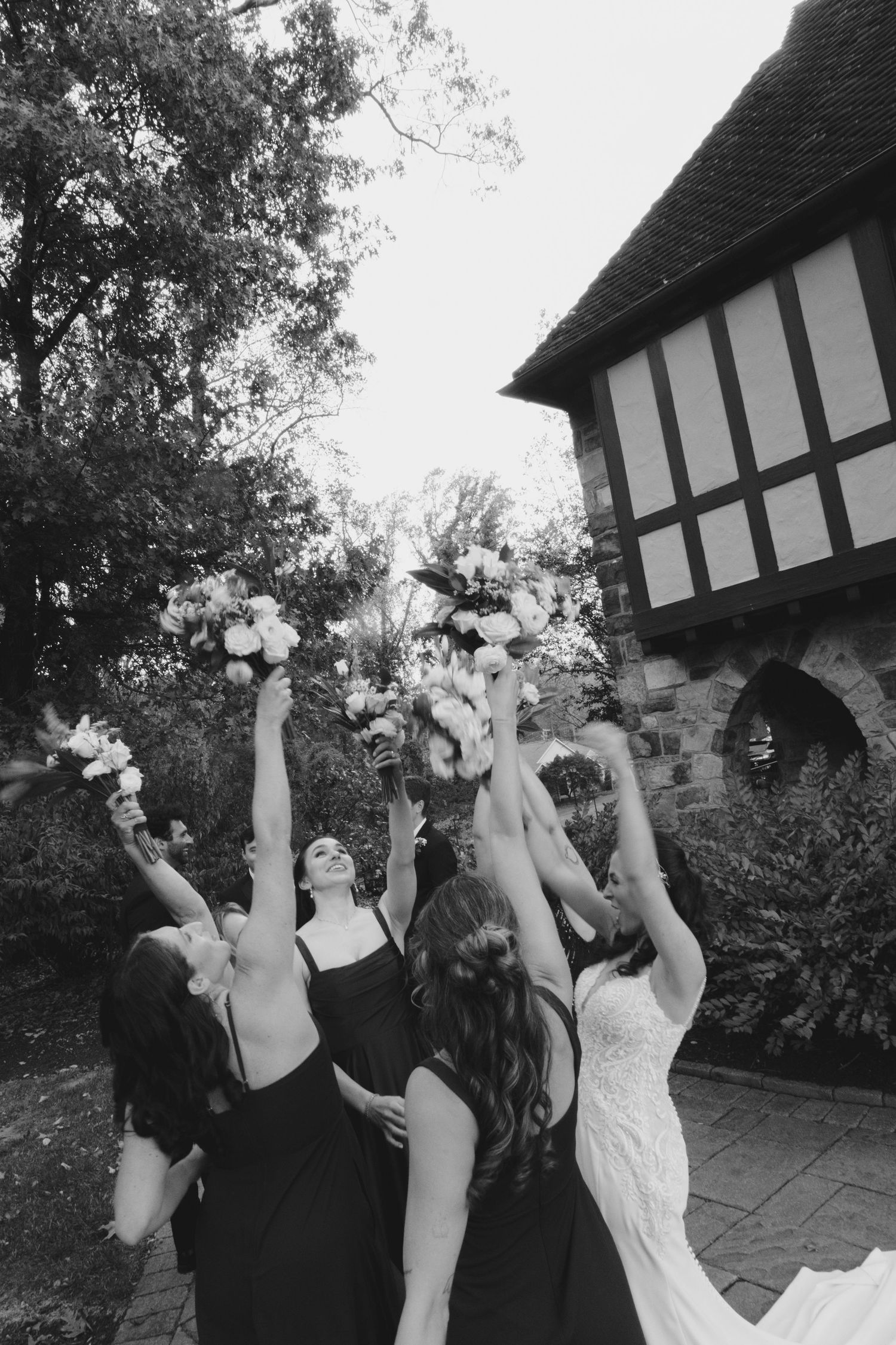 A group in formal attire throws flower petals into the air outside a Tudor-style building.