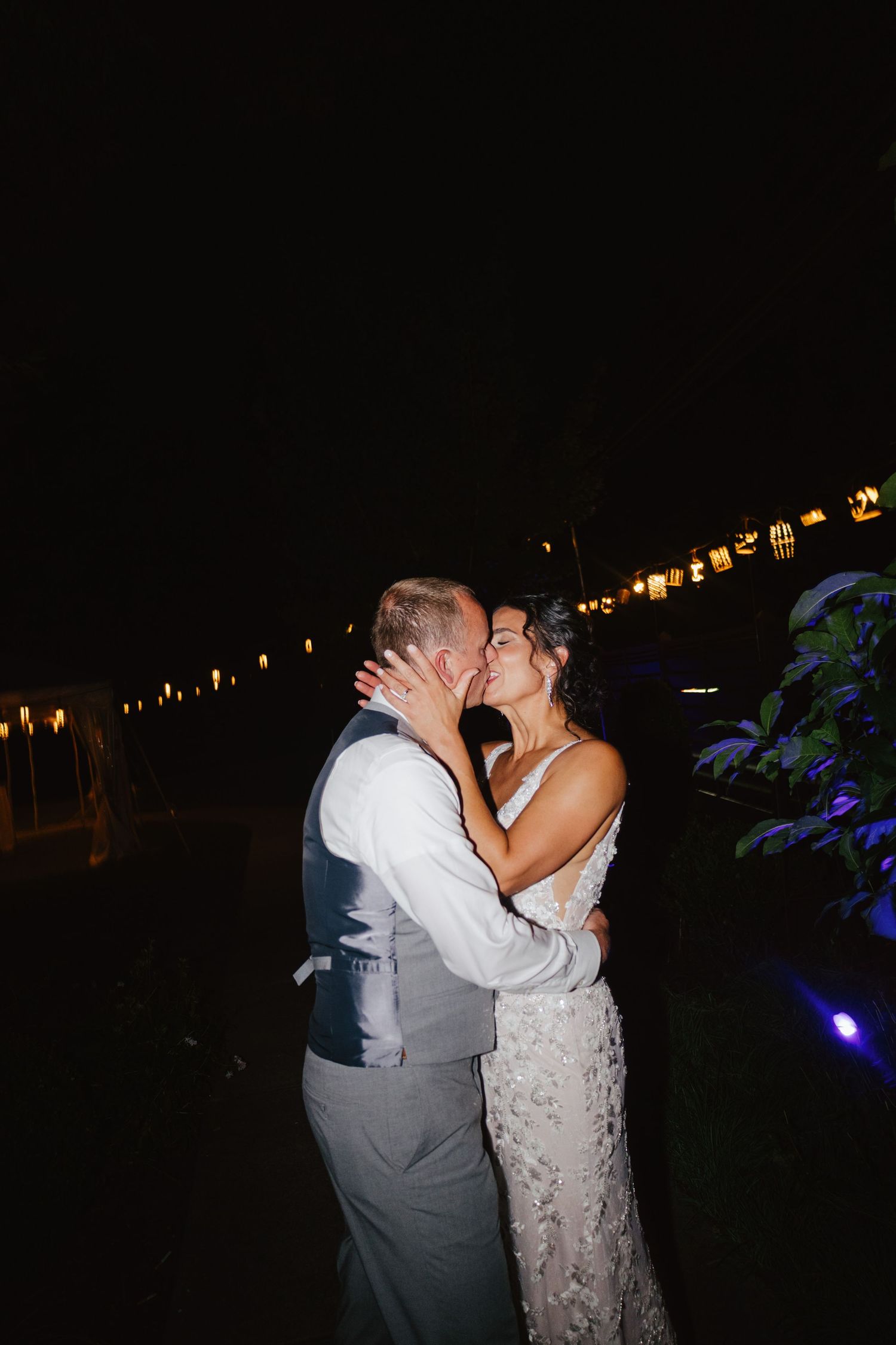 A couple shares a romantic kiss during their outdoor nighttime wedding reception with twinkling lights in the background.