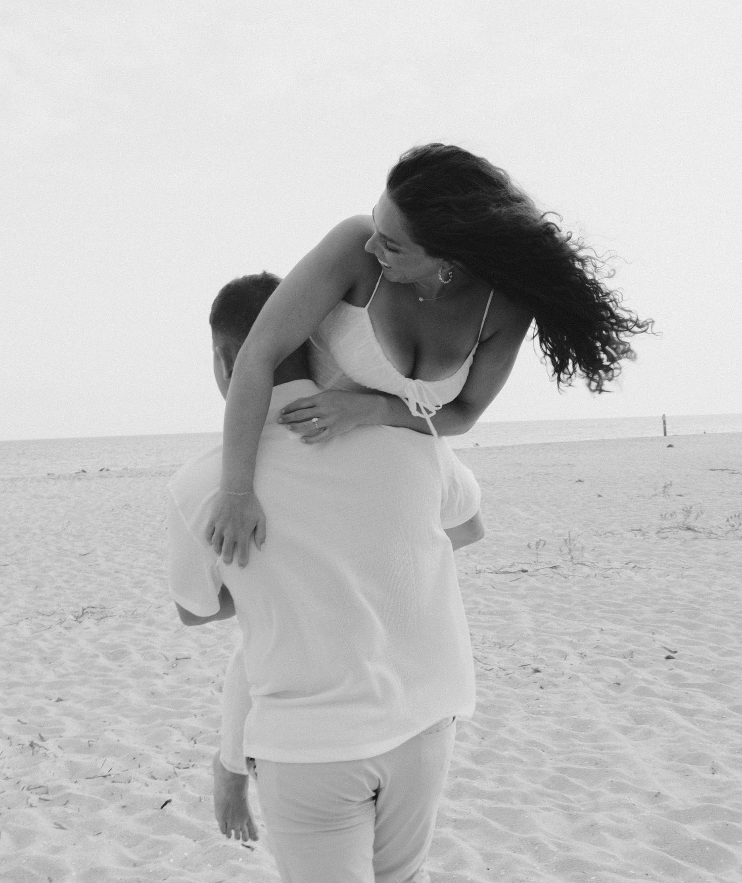 Black and white photo series of a couple dressed in white embracing playfully on a sandy beach.