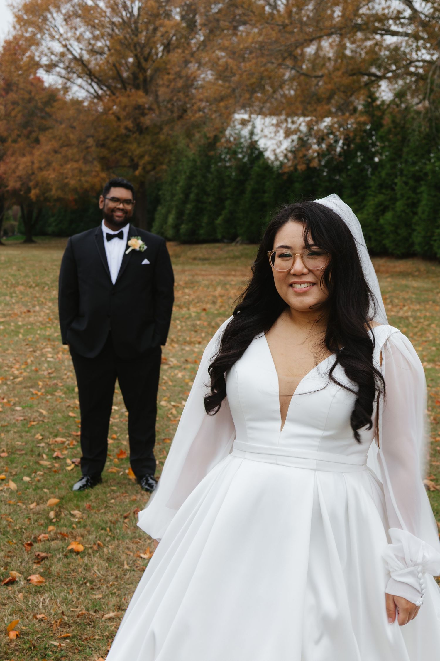 A couple poses for wedding photos on an autumn day with fallen leaves and evergreen trees in the background.