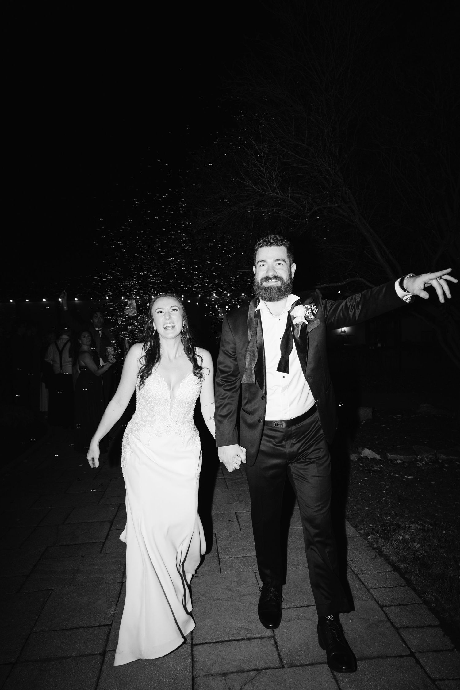 A couple walks together at night, with the person in white dress holding up their gown while walking on a paved path.