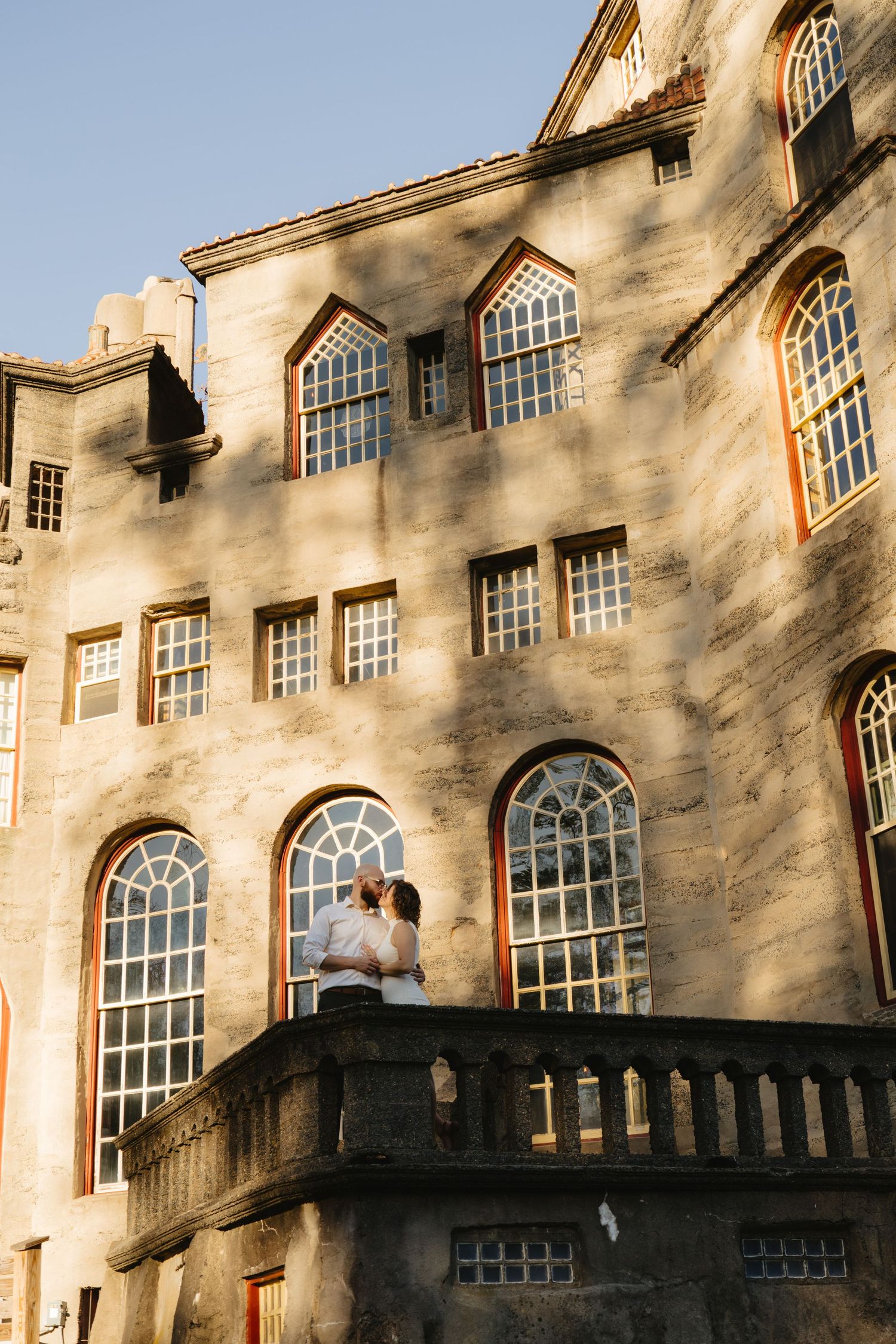 A romantic couple embraces on a stone balcony of a historic building with arched windows and gothic architecture.