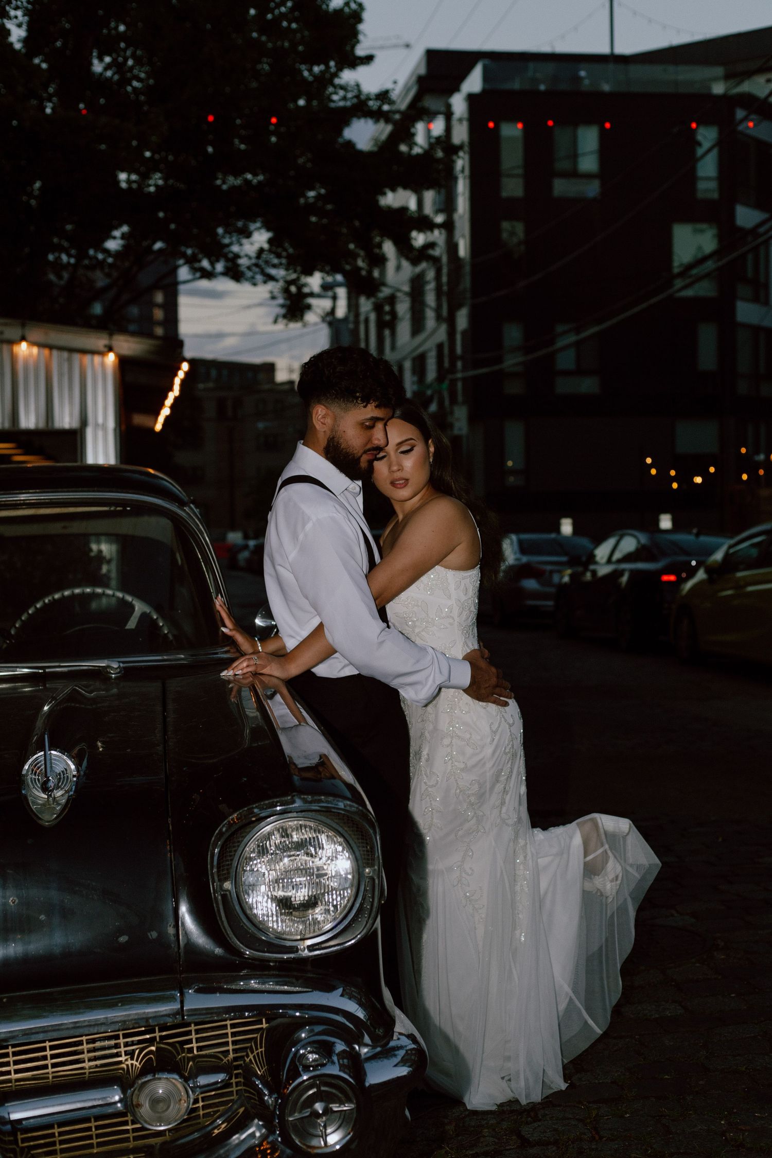 Romantic couple pose against vintage car during evening wedding photoshoot.