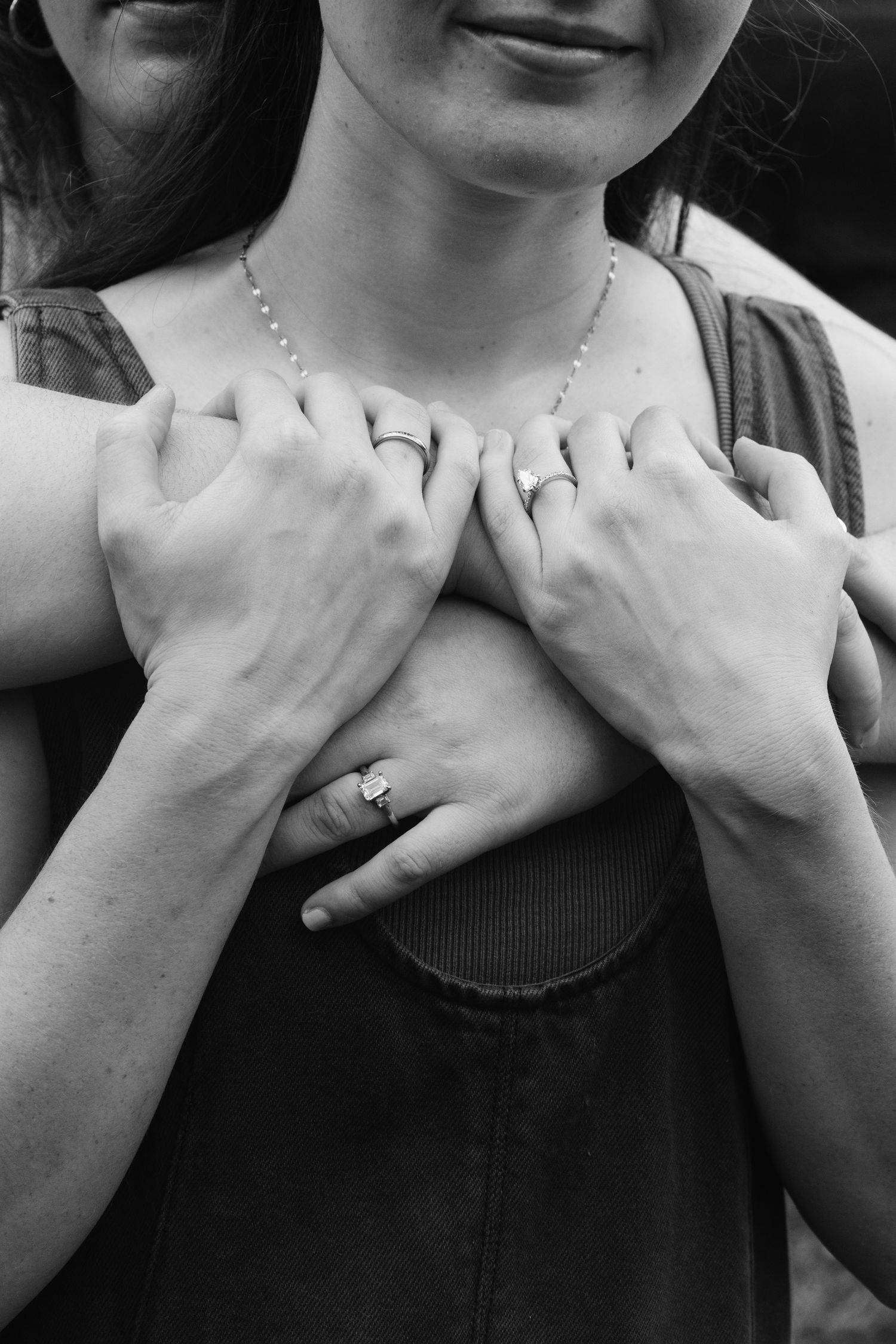 Black and white close-up of hands embracing from behind, with an engagement ring visible on one finger.