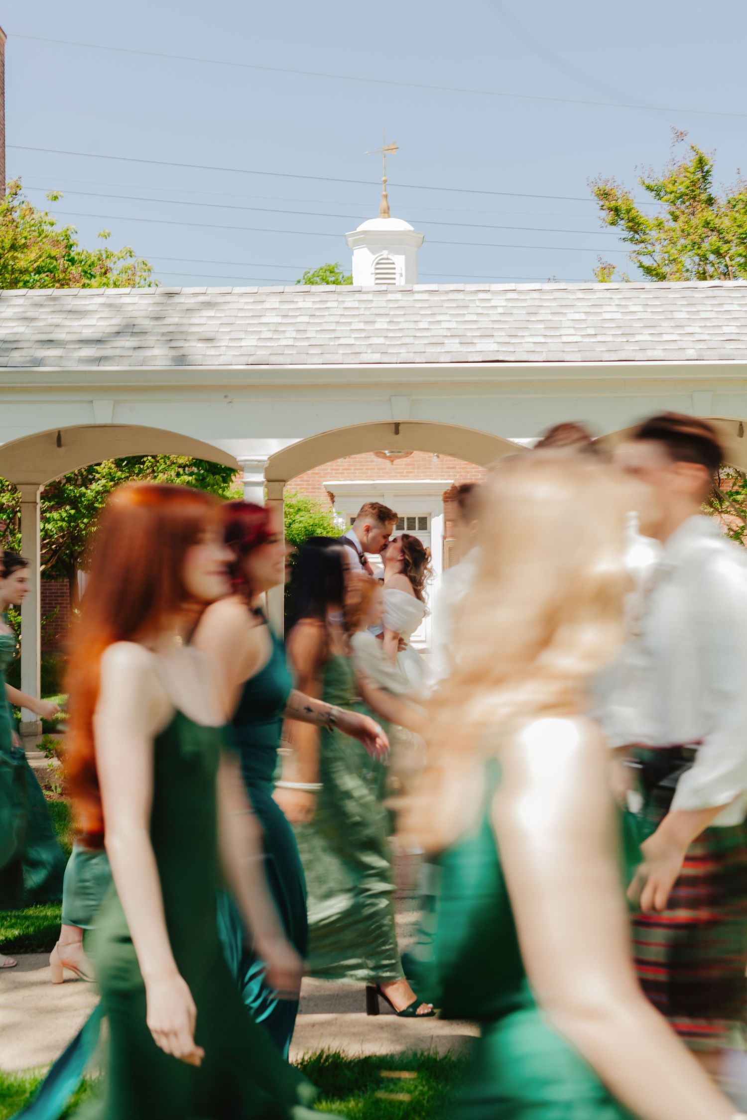 Motion-blurred wedding celebration with guests dancing in emerald dresses under a white gazebo.