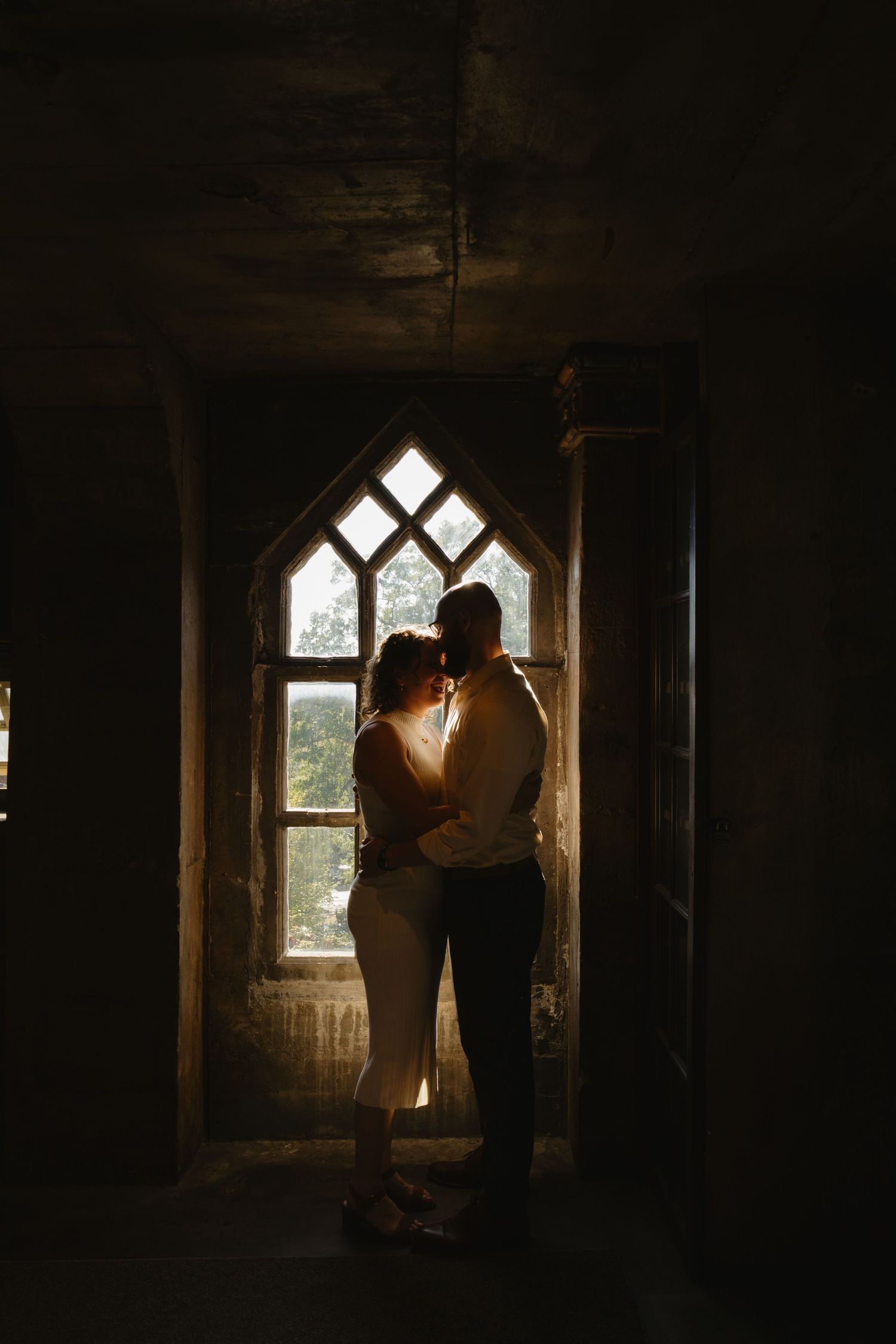 Two silhouettes stand together in front of a gothic window frame in a dark room with dramatic lighting.