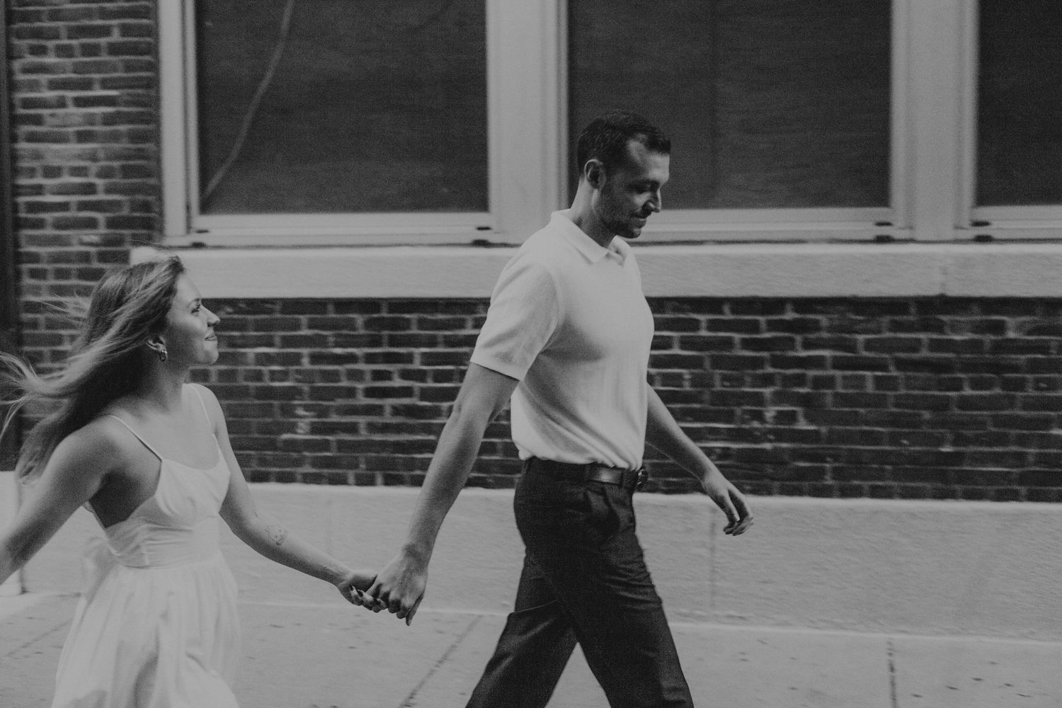 A couple walking together on a sidewalk in black and white, with the woman wearing a flowing dress.