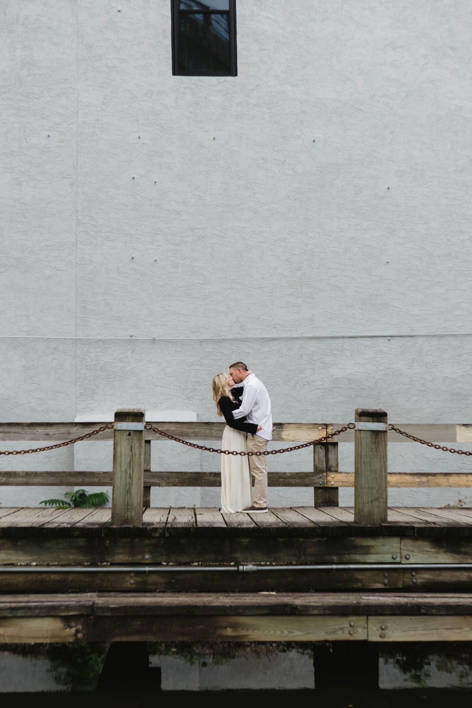 Two figures sit on a wooden boardwalk railing overlooking water on an overcast day.